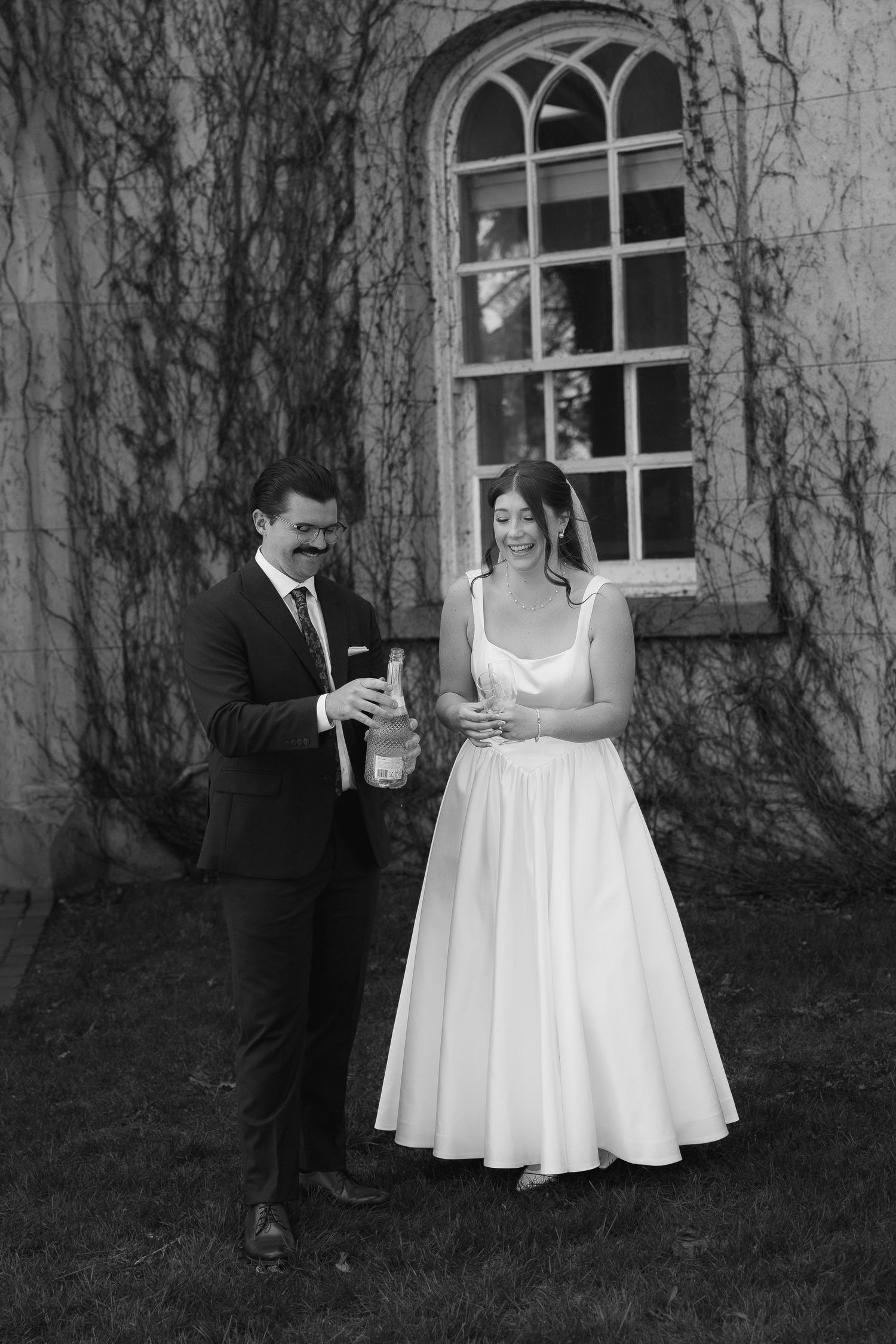 Black and white photo of a bride and groom opening champagne outside a ivy-covered building on their wedding day.