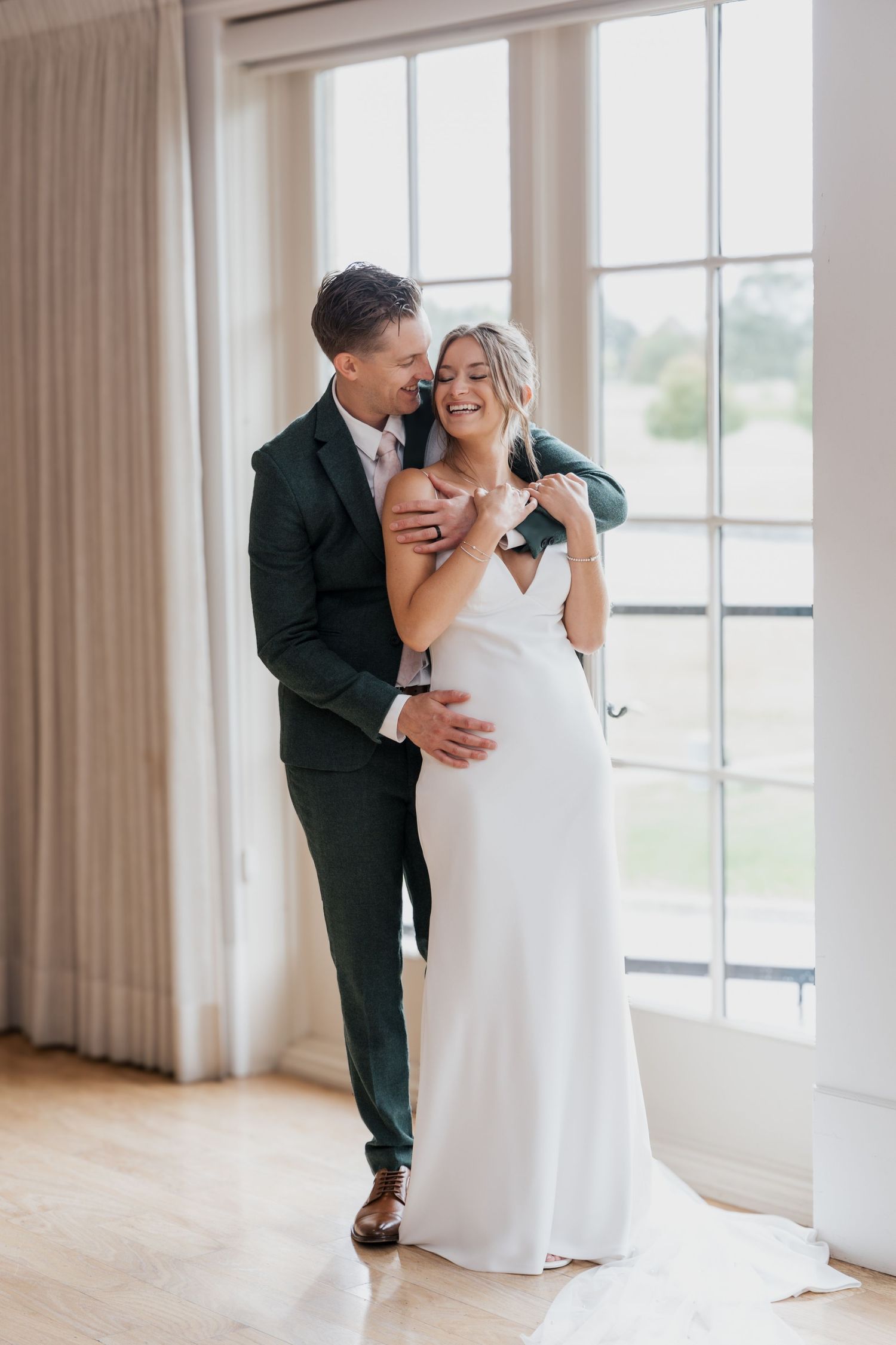 A groom in a dark suit embraces his smiling bride in a white gown near large windows in an elegant venue.