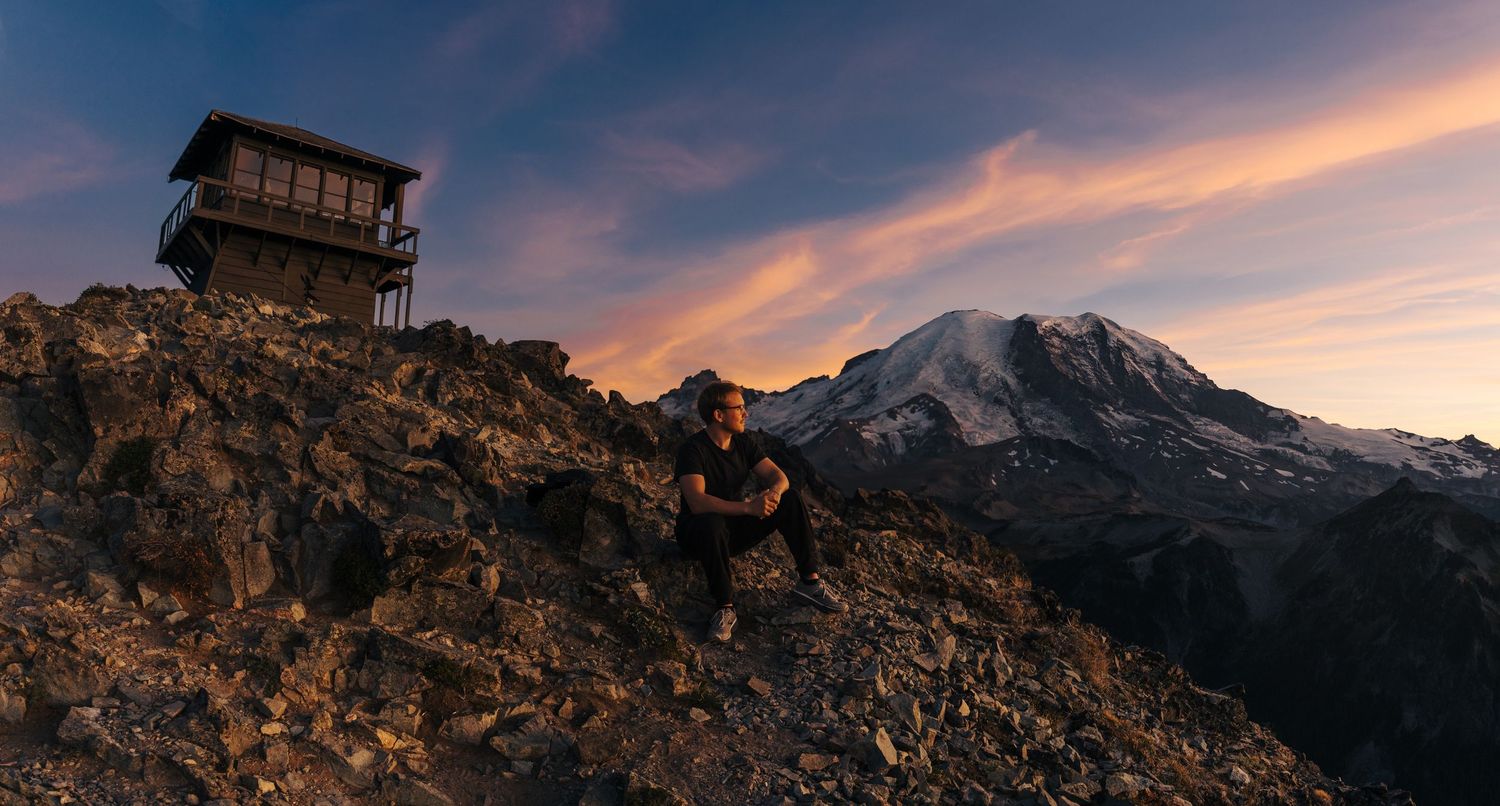 Hiker resting on rocky summit near a fire lookout tower at sunset with Mount Rainier in the background.