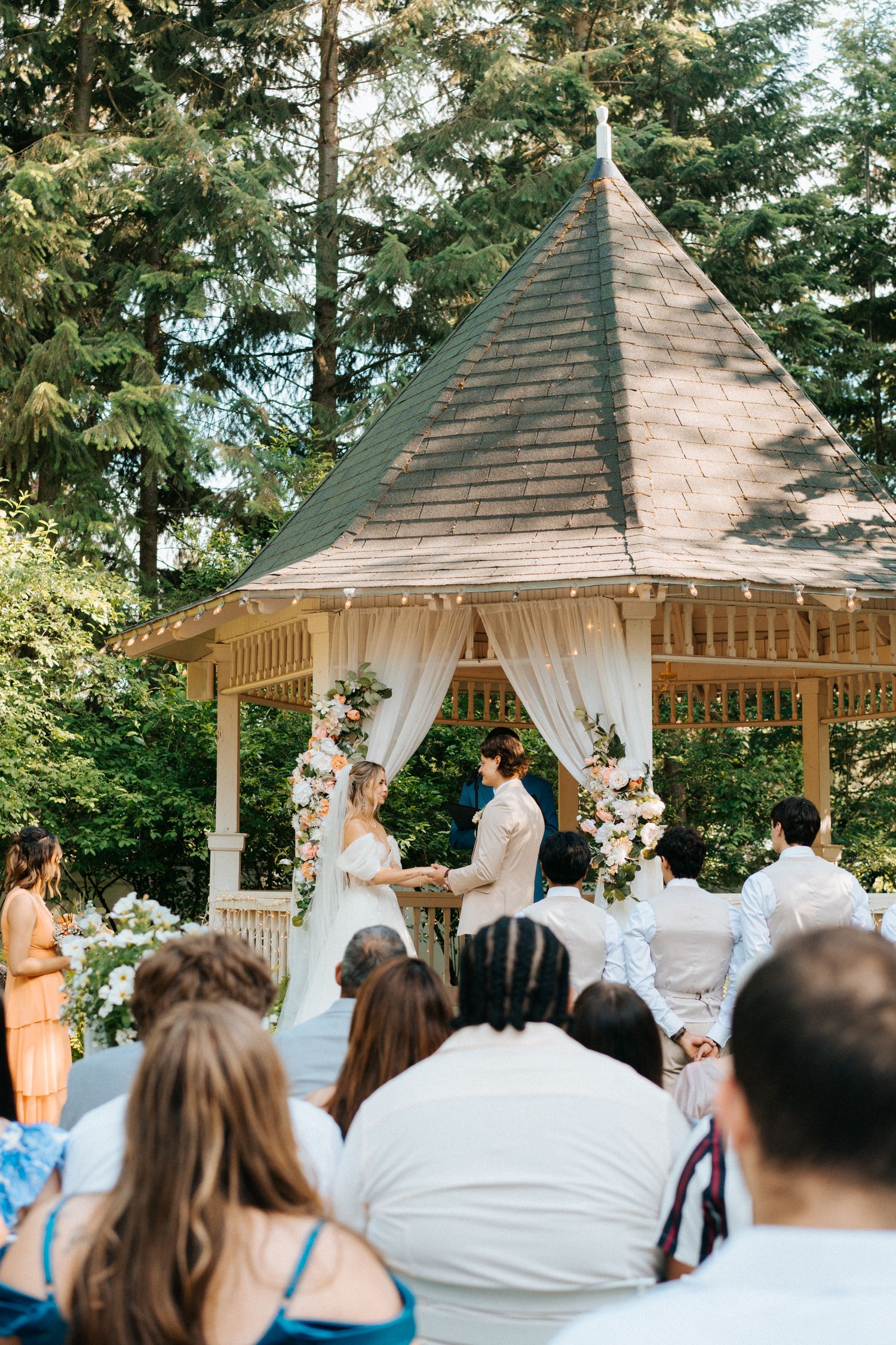Wedding ceremony taking place under a white-draped wooden gazebo surrounded by trees and guests.