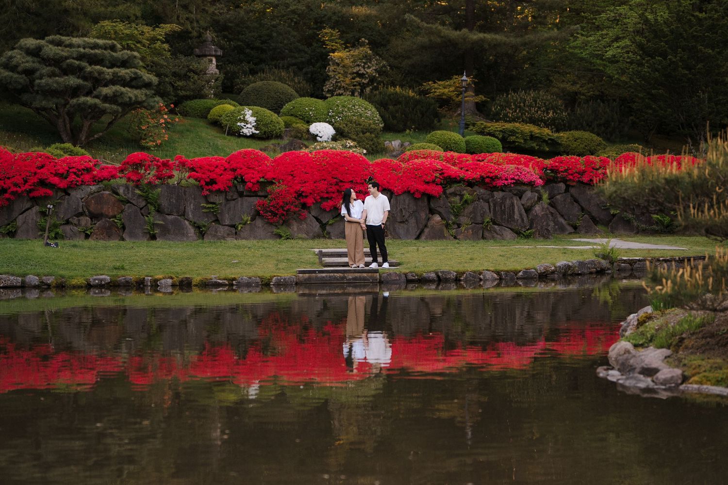 Couple standing on a dock by a tranquil pond surrounded by vibrant red azalea bushes in a Japanese garden.