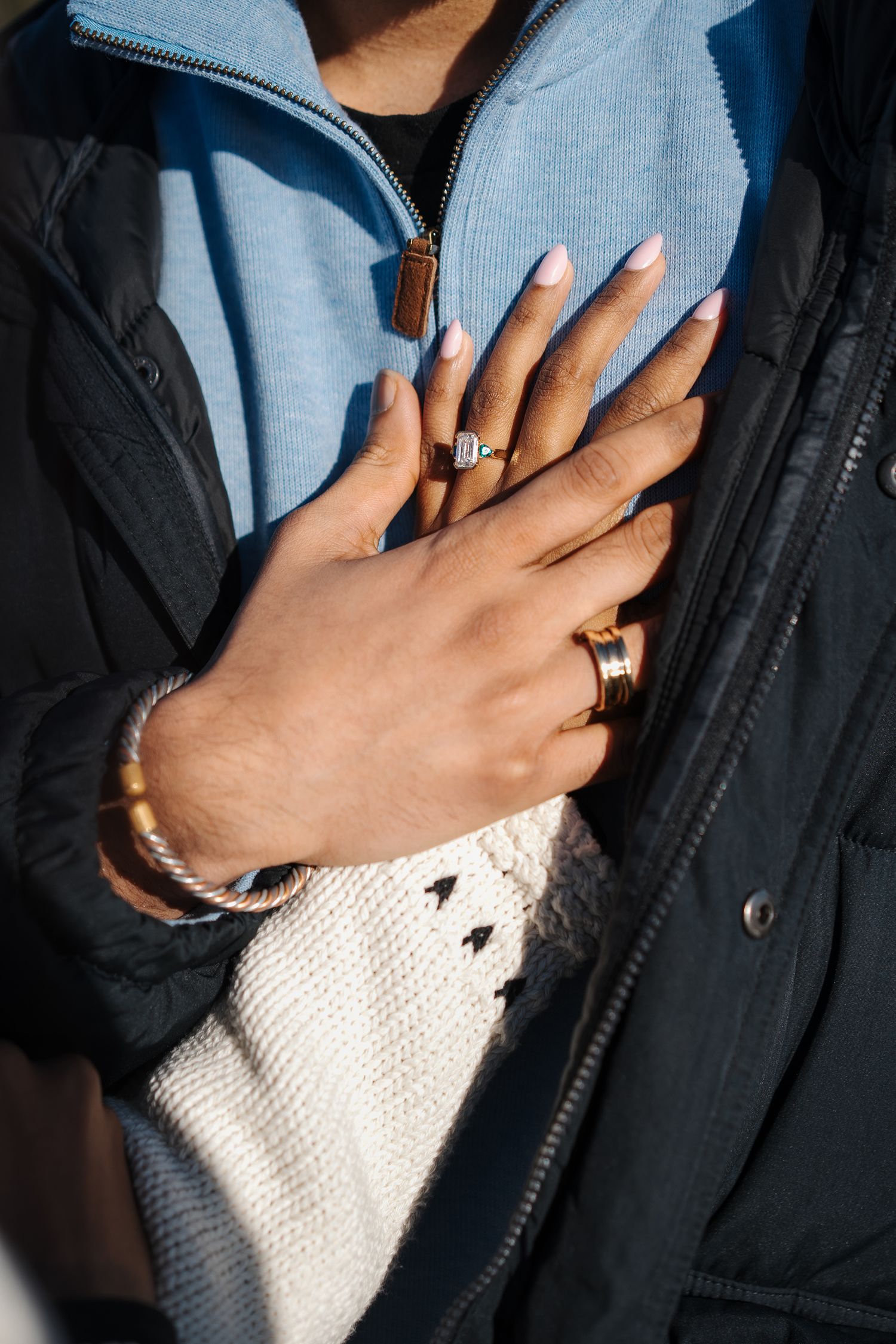 Close-up view of hands wearing gold rings and bracelets against navy vest and white knit sweater in natural light.