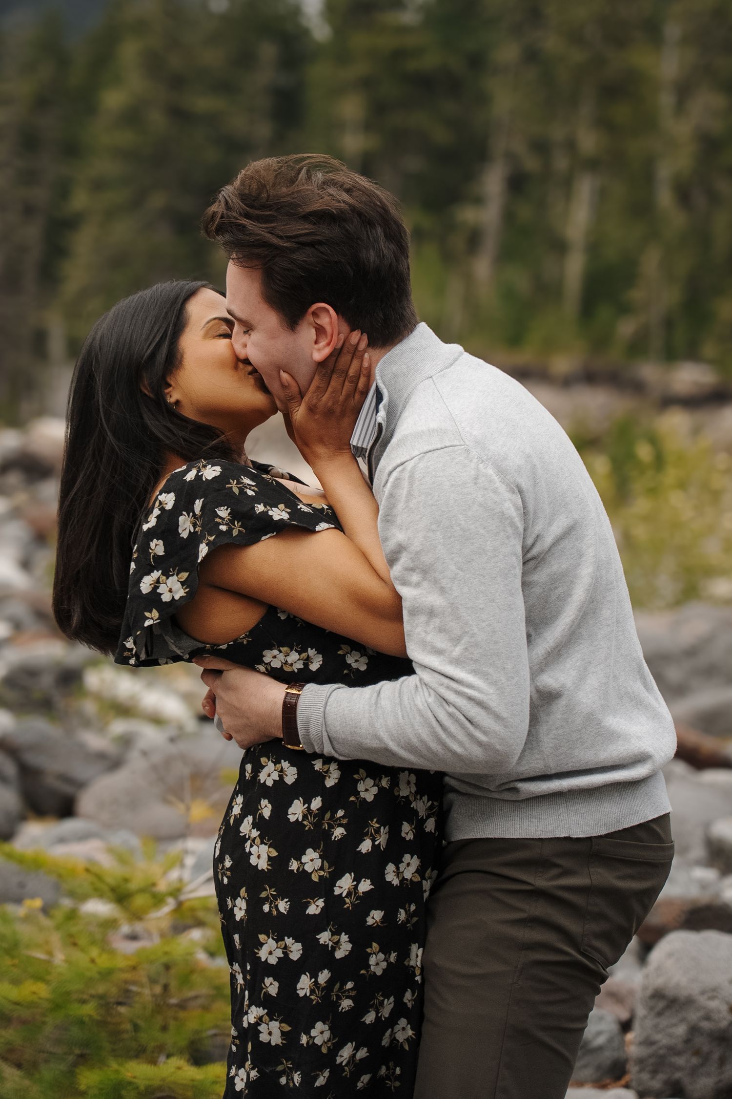 A romantic couple embraces and shares a tender kiss outdoors in a natural setting with rocks and trees in the background.