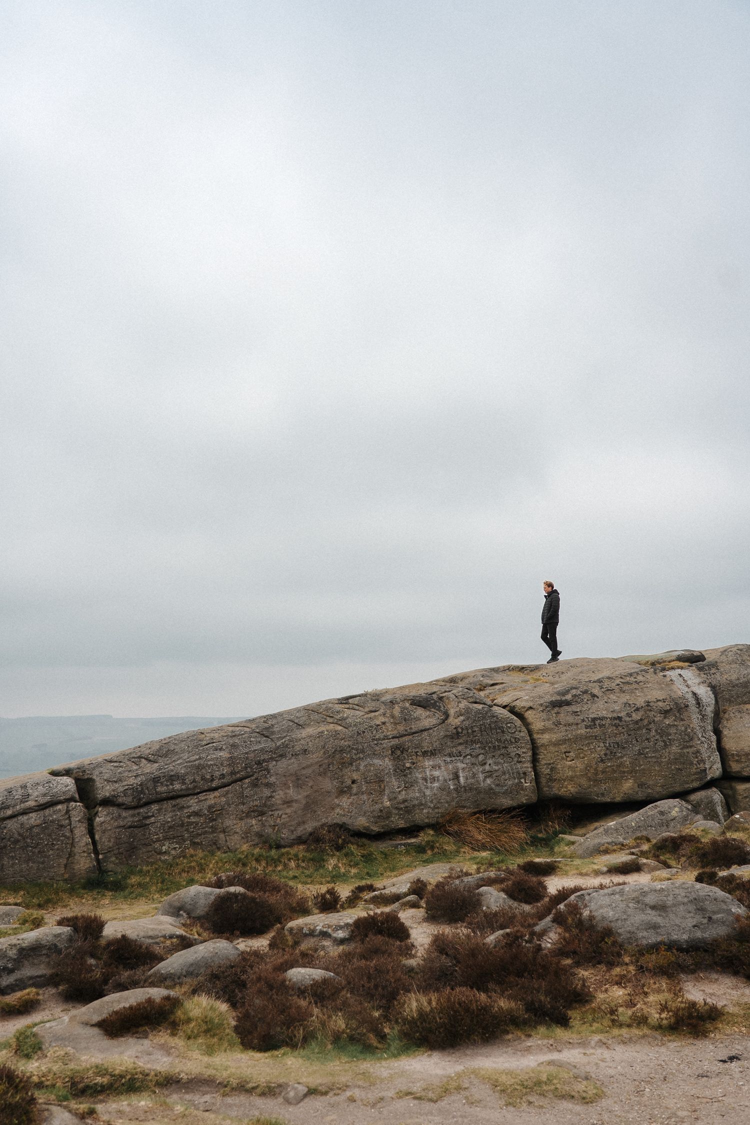 Person standing atop a large granite rock formation on an overcast day with expansive moorland views.