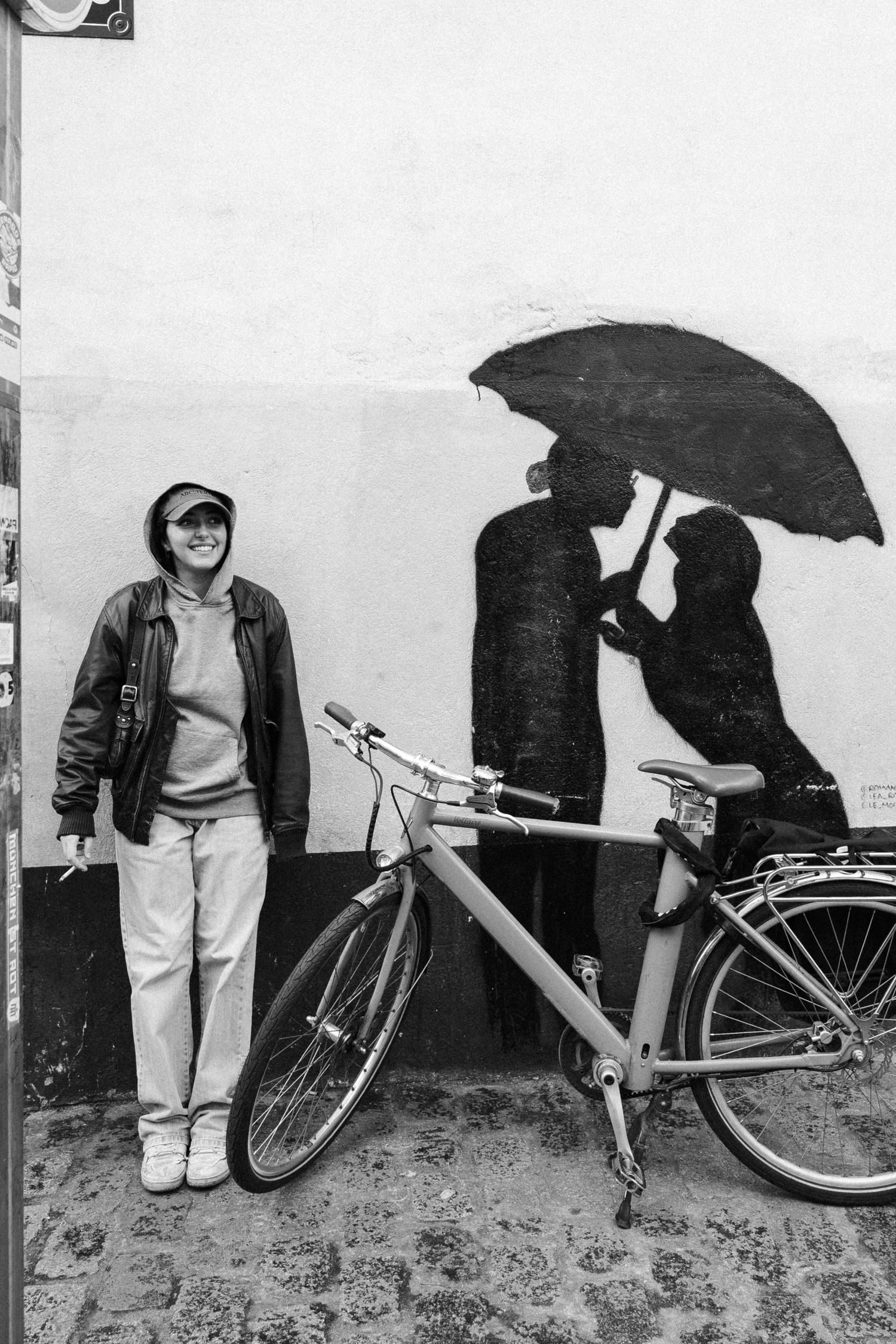 Black and white photo of a smiling person in a jacket standing beside a bicycle against a wall with couple silhouette graffiti.