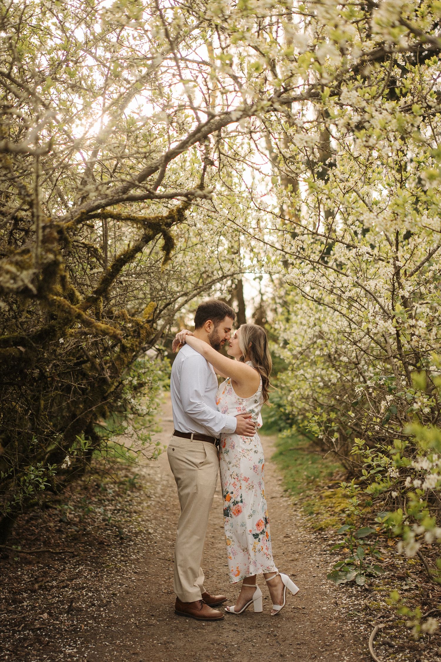 A couple shares a romantic moment on a sunlit path surrounded by blooming spring trees and white flowers.