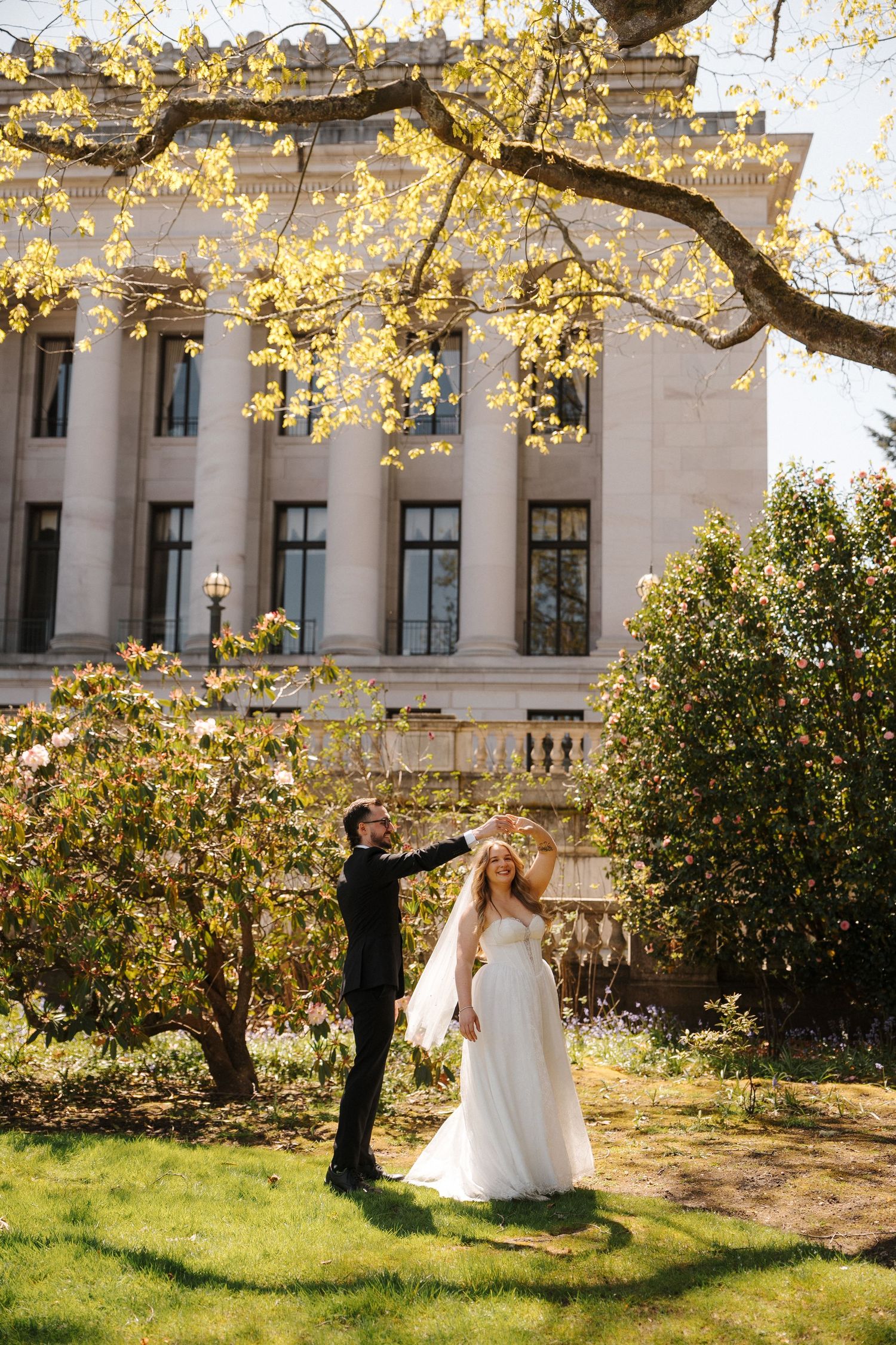 Groom lifts bride's veil under blooming tree outside elegant neoclassical building during spring wedding ceremony.