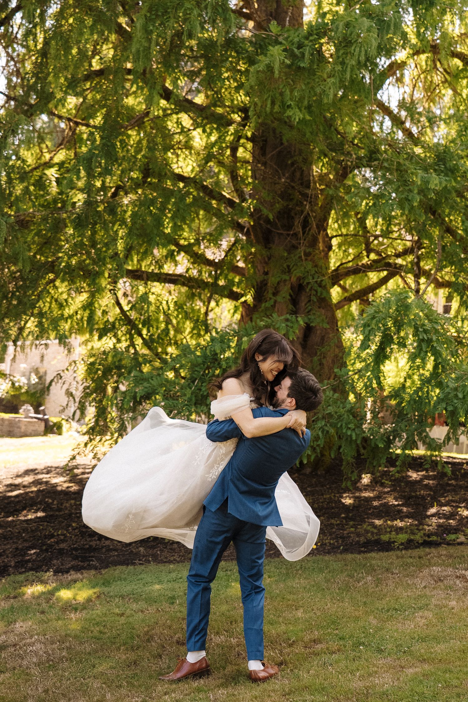A romantic couple embraces under a large willow tree in a park setting with soft natural lighting.