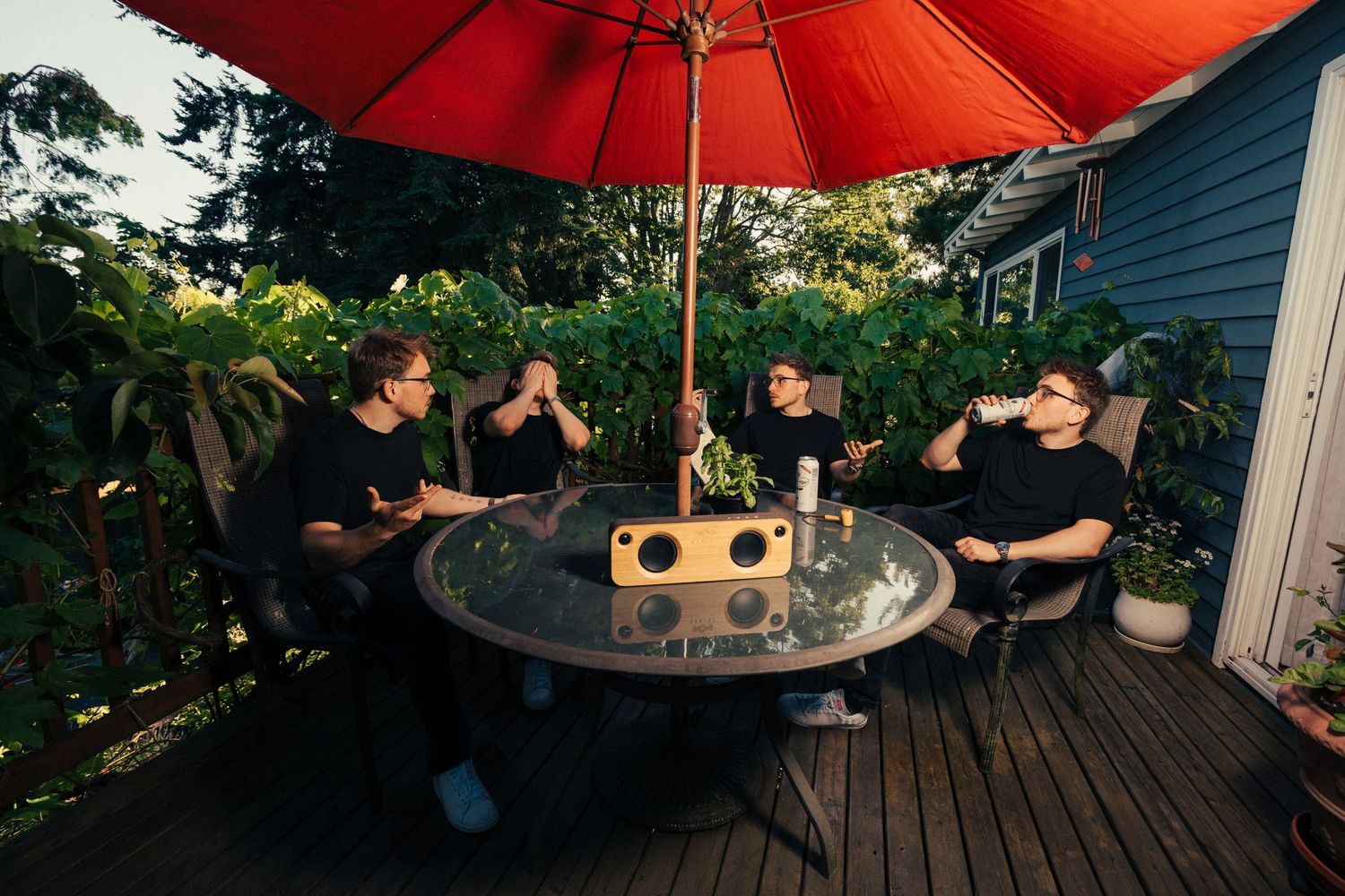 Four friends sitting at an outdoor patio table under a red umbrella with a Bluetooth speaker centerpiece.
