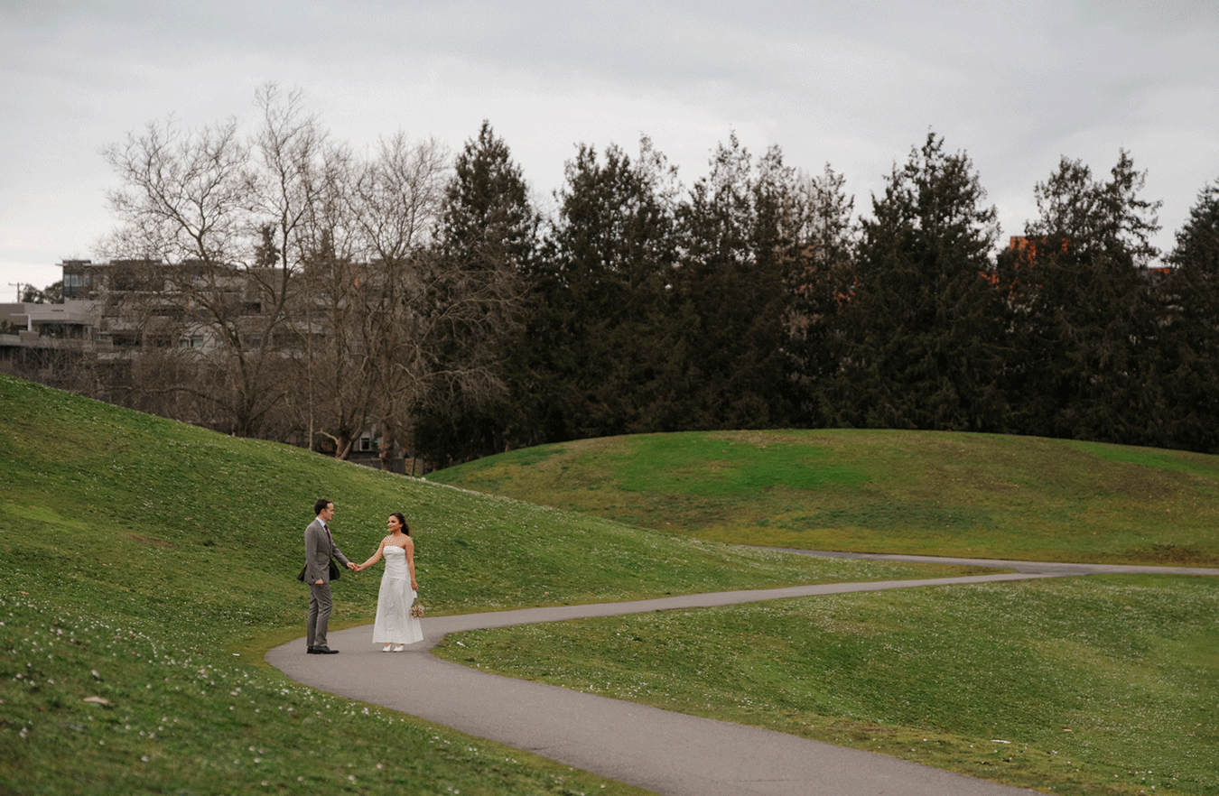 Bride and groom holding hands on a winding path in a lush green park with trees and a building backdrop.