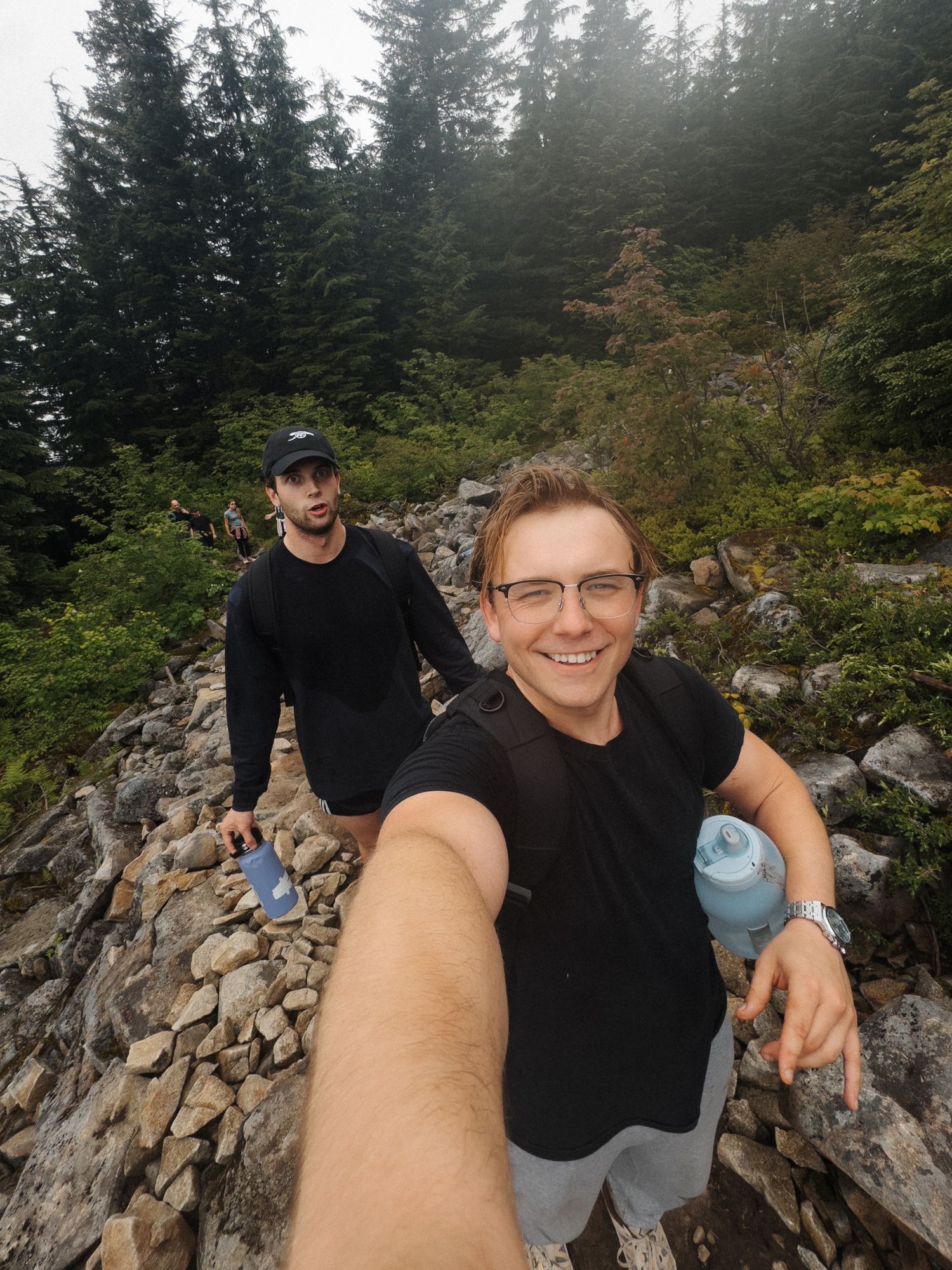 Two hikers smiling on a rocky trail surrounded by misty evergreen forest, one taking a selfie holding a water bottle.