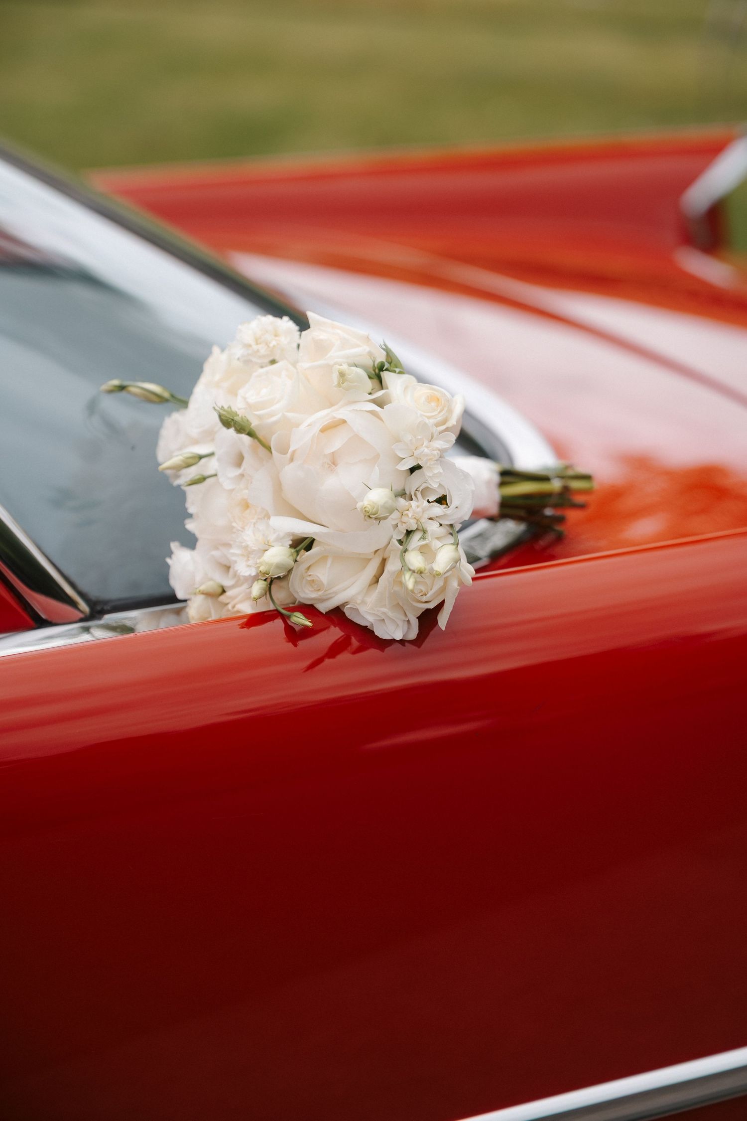 White bridal bouquet of roses and peonies resting on a shiny red classic car hood.
