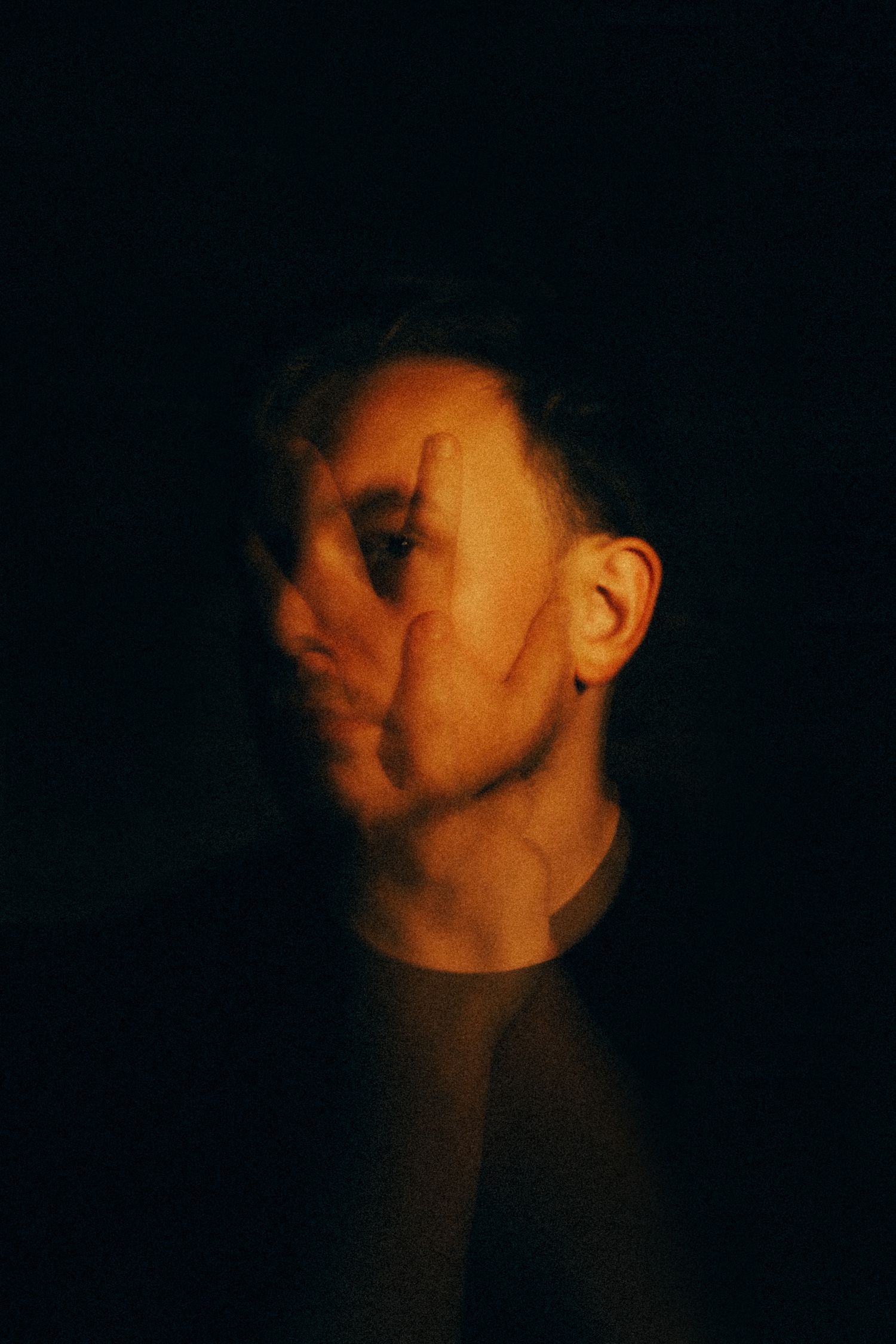 A person partially covering their face with their hand in dramatic low-key lighting against a dark background.