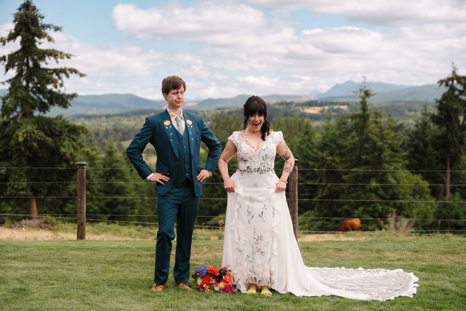 Bride and groom pose outdoors at a Pacific Northwest wedding venue with mountain views, she in lace gown, he in teal suit.