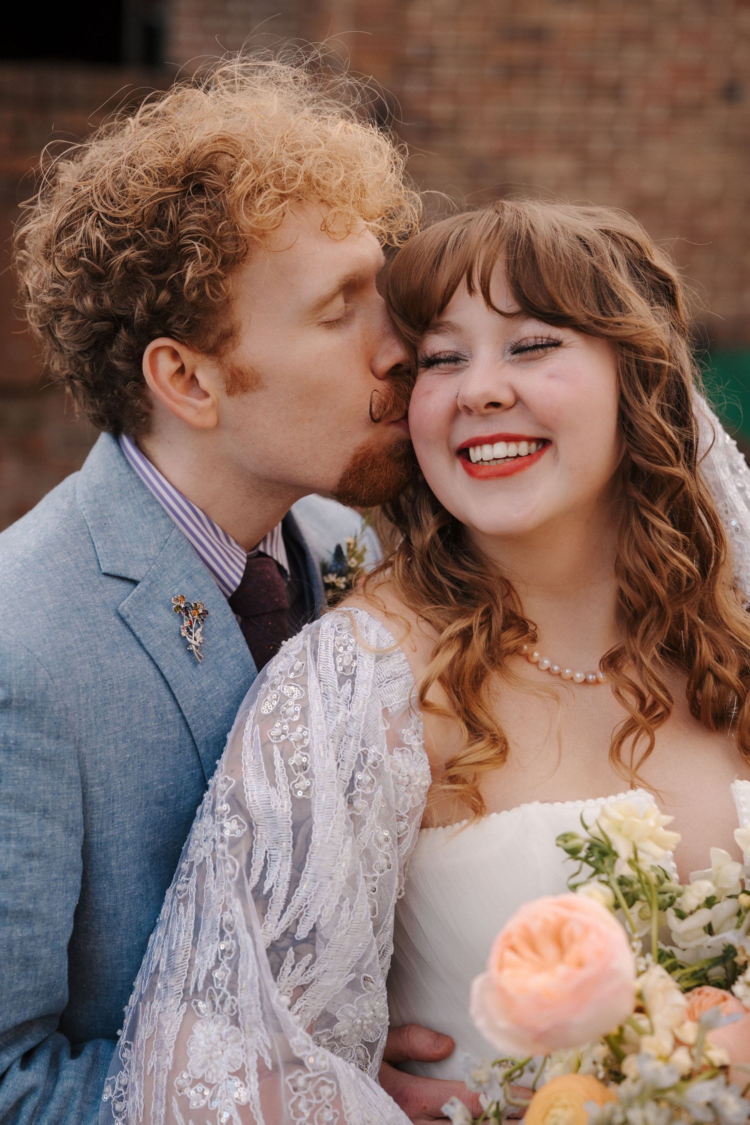 A groom with red curly hair kisses his smiling bride holding a pastel bouquet on their wedding day.