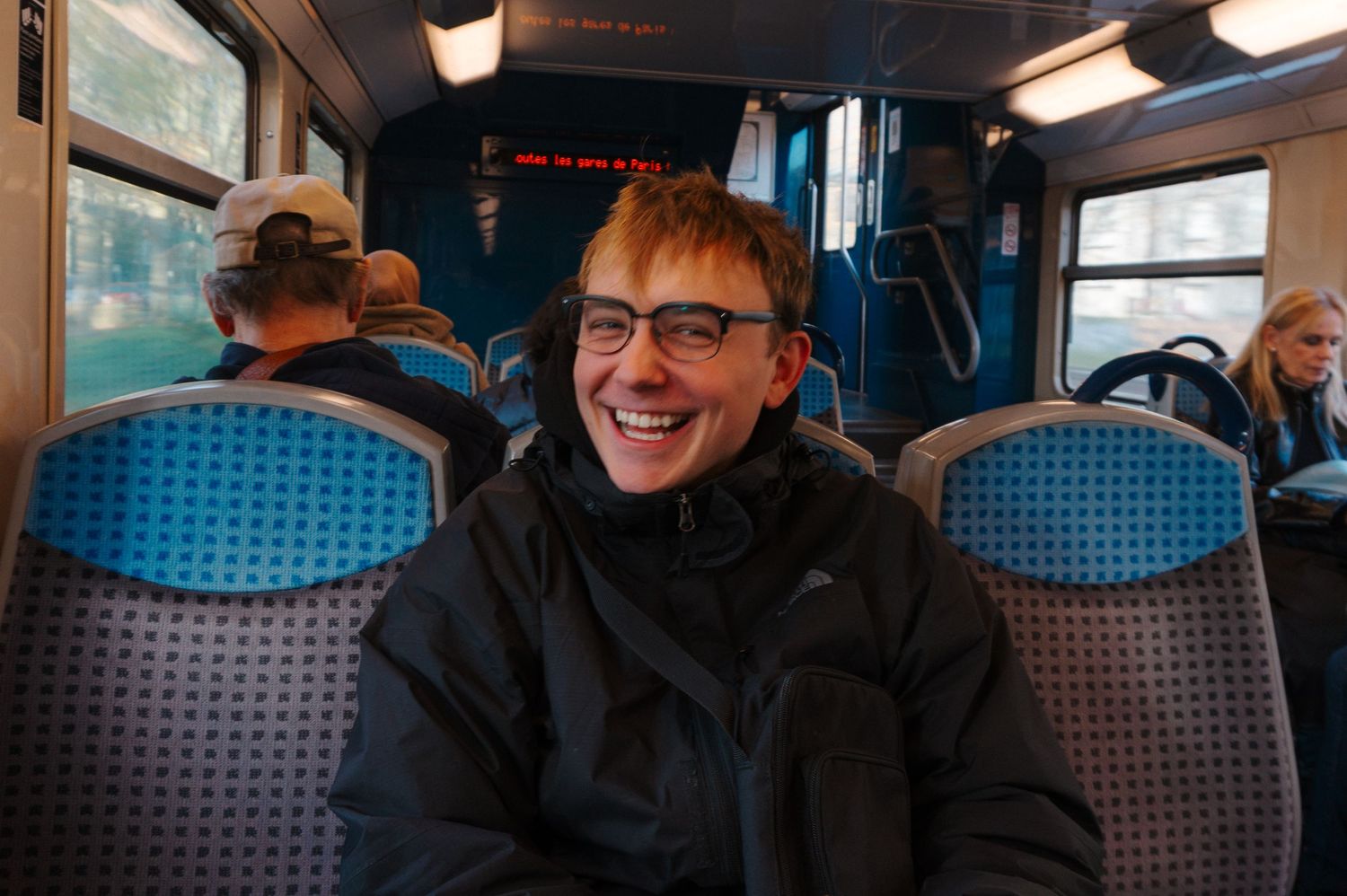 Smiling young person with red hair and glasses sitting on a bus with blue seats.