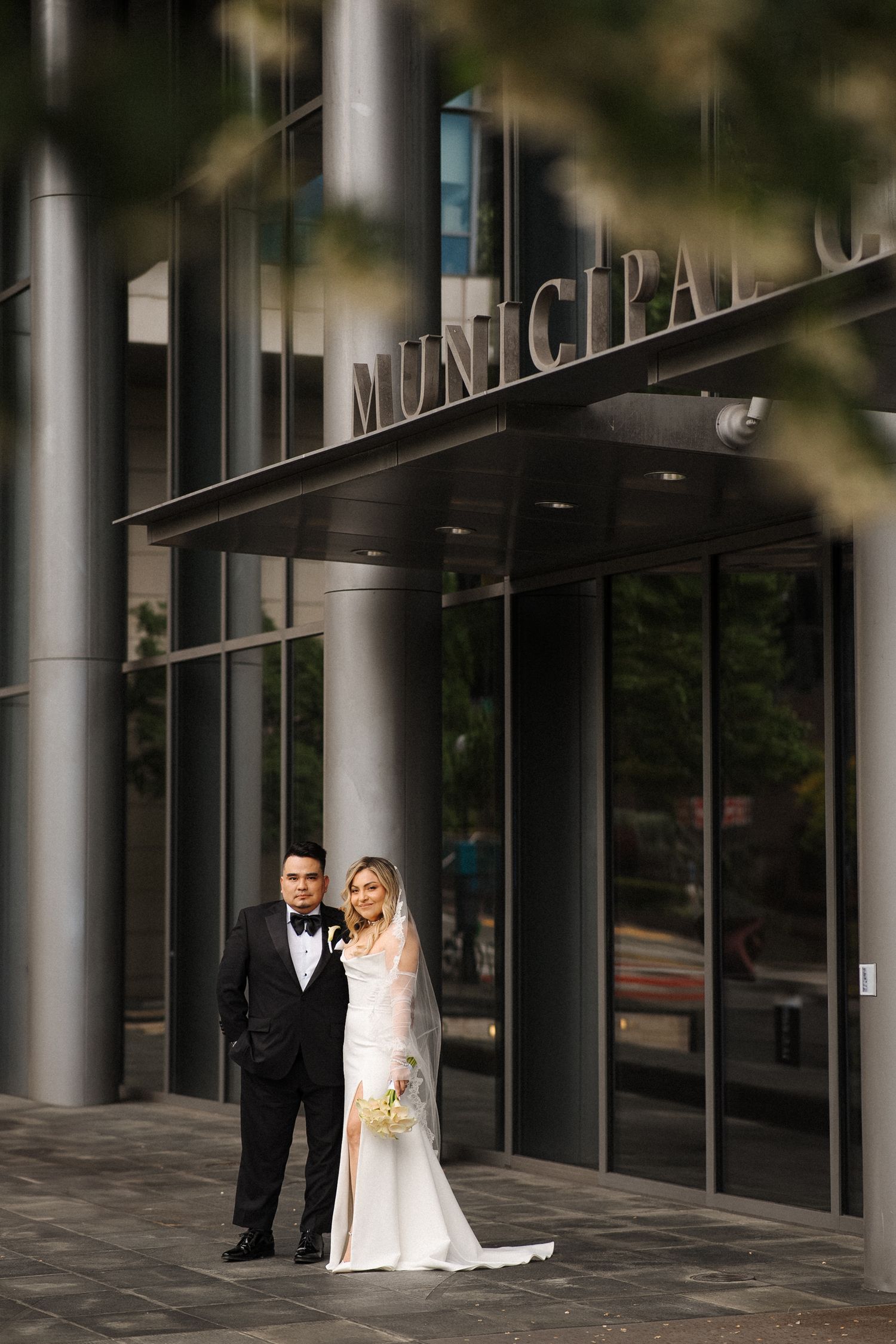 A bride and groom share a romantic moment outside a modern glass building with pillars.