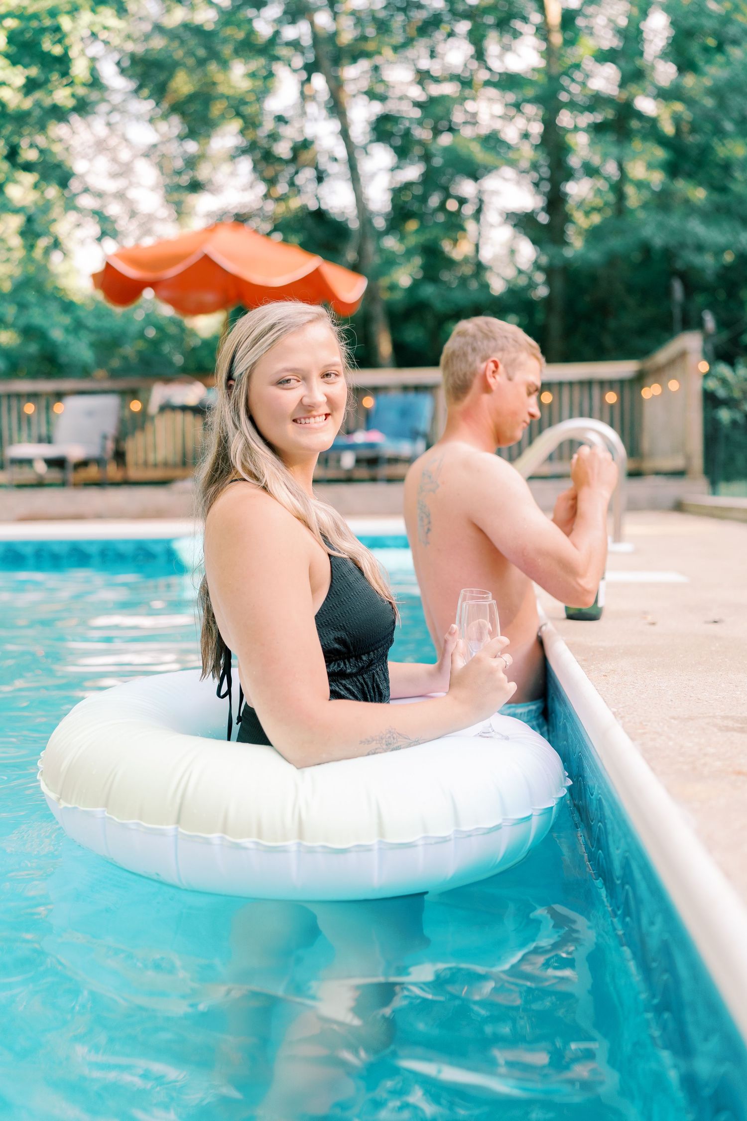 Two people relax on white pool floats in a backyard swimming pool.