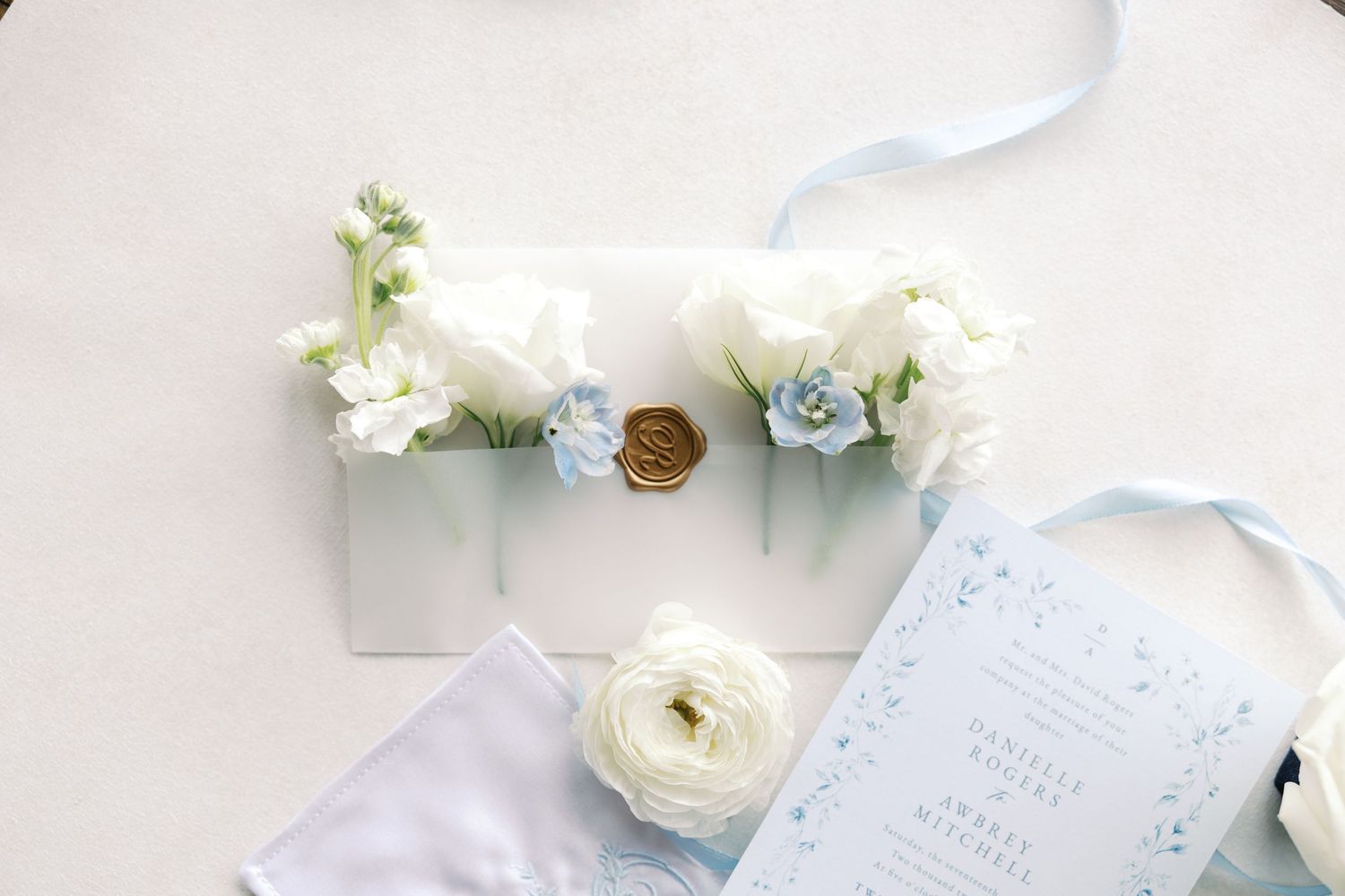 Wedding details featuring white flowers, blue ribbon, and elegant stationery on light background.