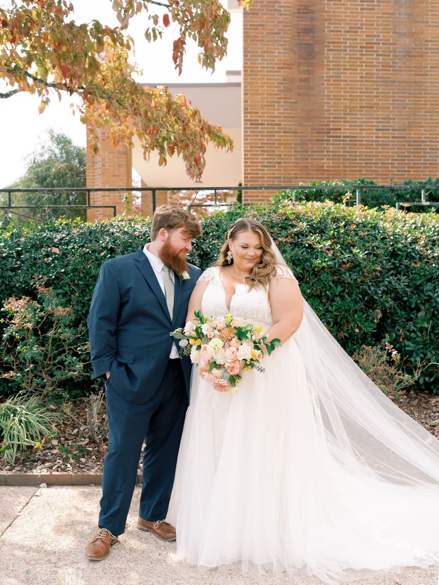 A wedding couple poses outdoors with a flowing white dress train and peach bouquet.