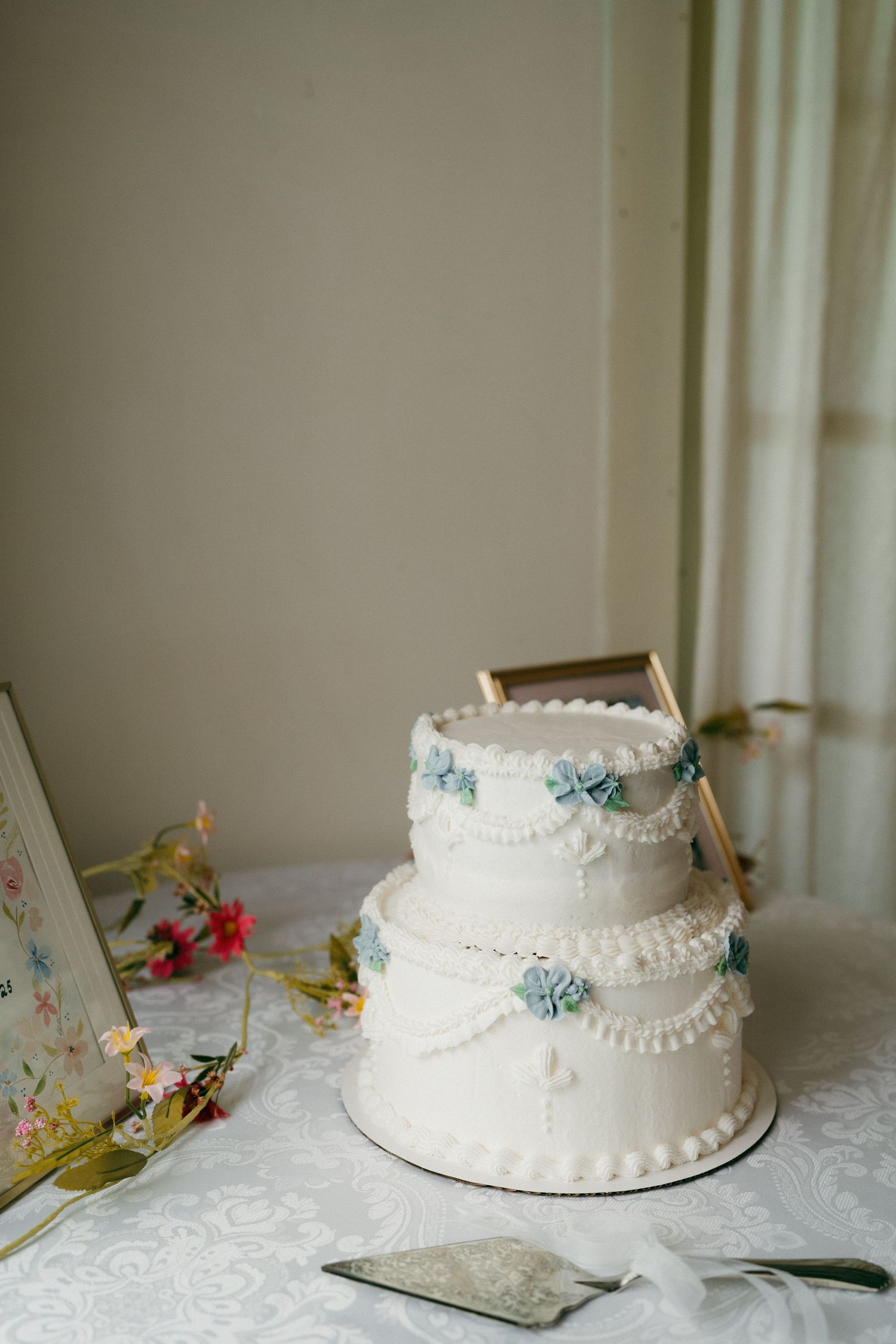 White two-tiered wedding cake decorated with delicate pearls and blue floral accents on a lace tablecloth.