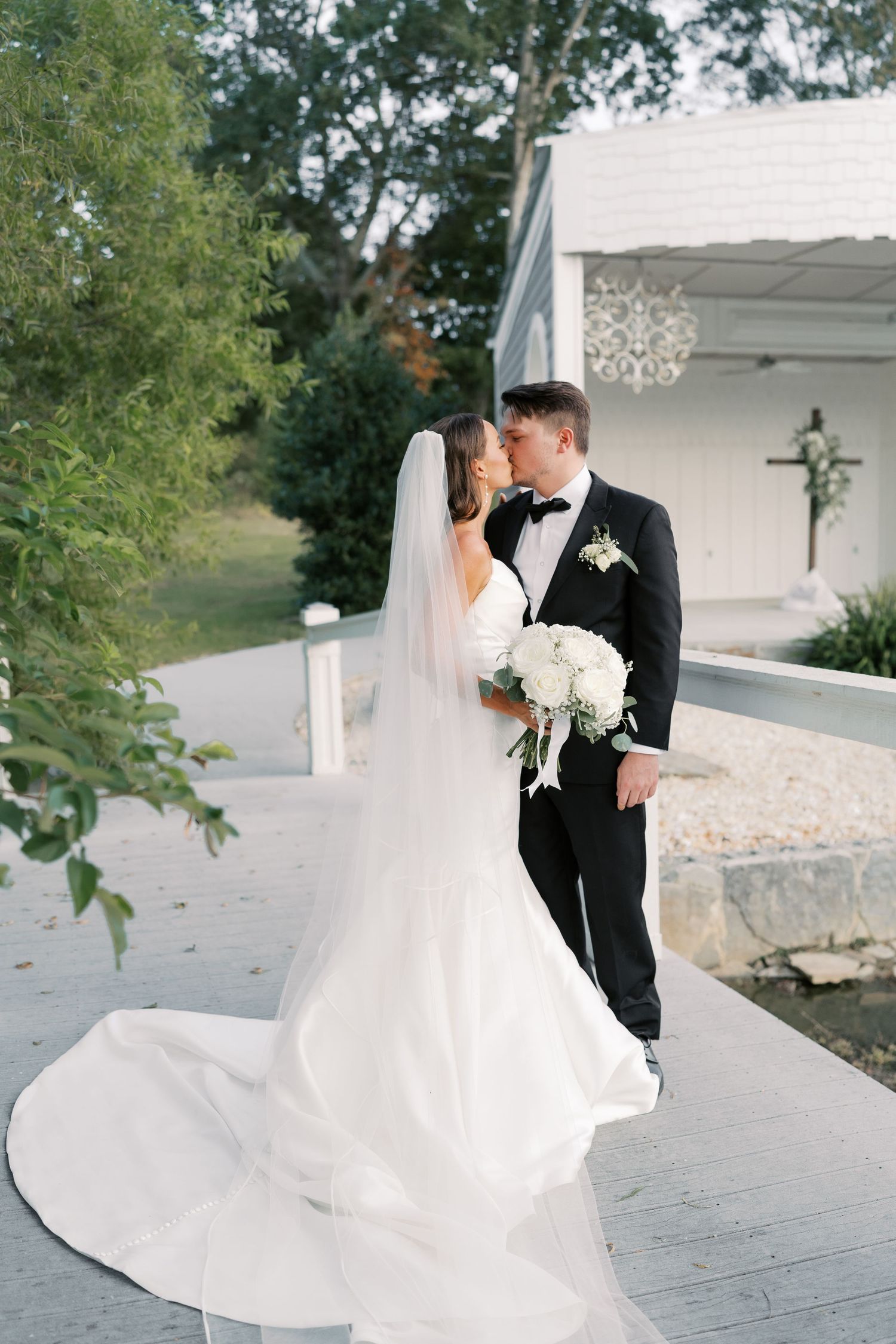 A romantic wedding couple sharing a kiss on a white porch with a flowing bridal gown and bouquet.