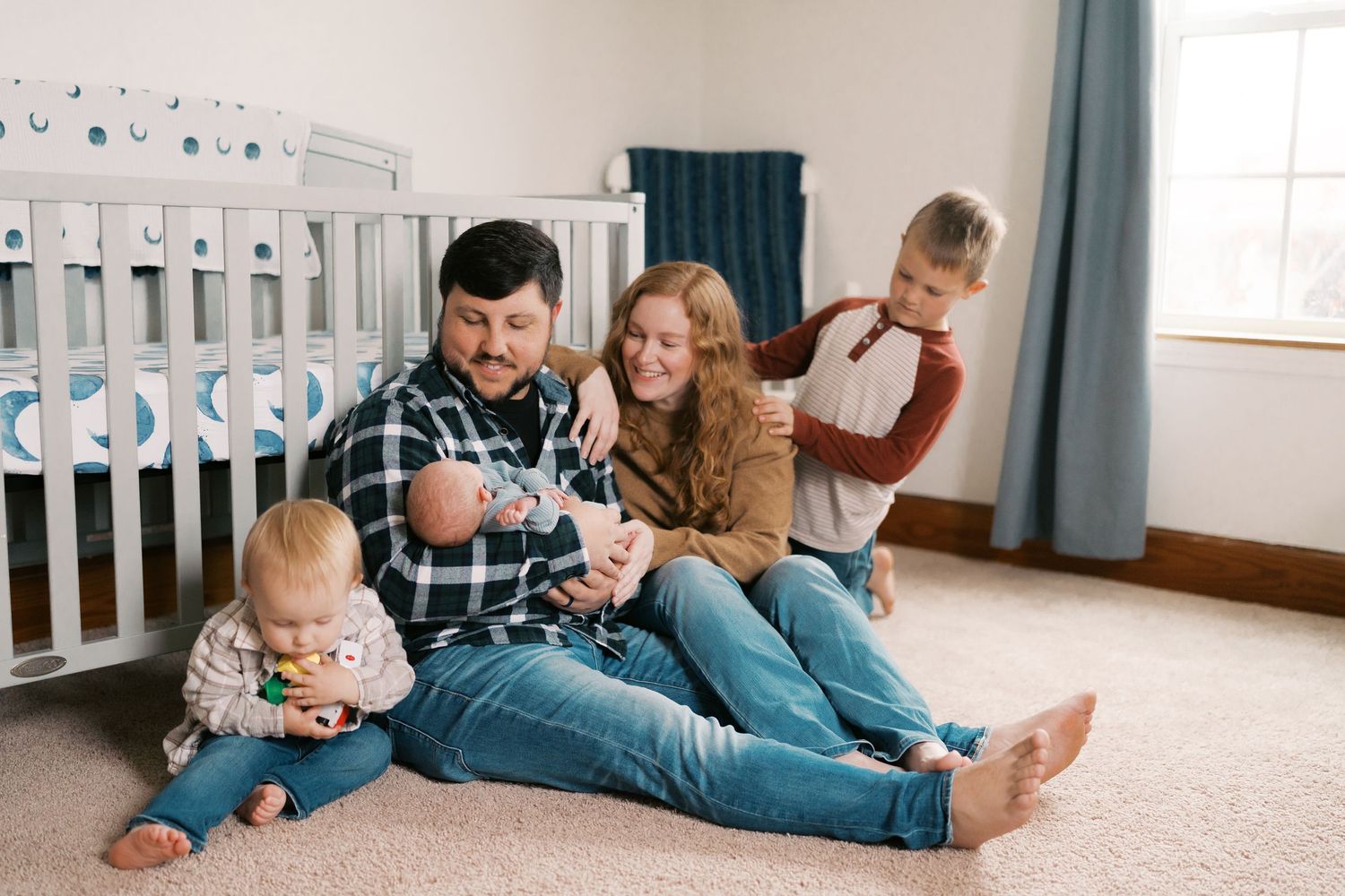 Interior view of a nursery with white crib, blue curtains, and family members playing on carpet.