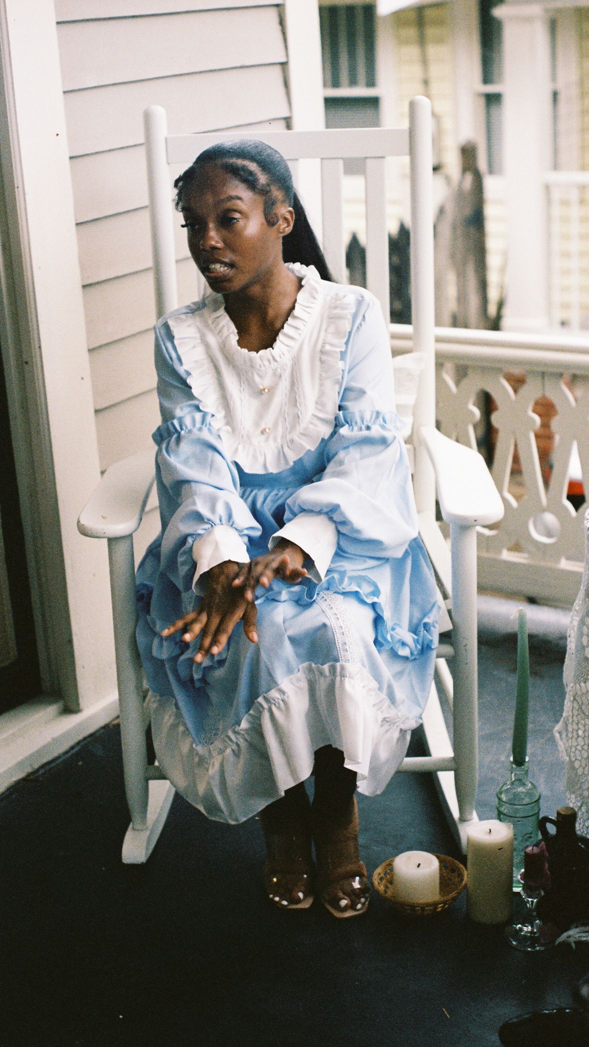 Person in a light blue and white dress sitting in a rocking chair on a porch.