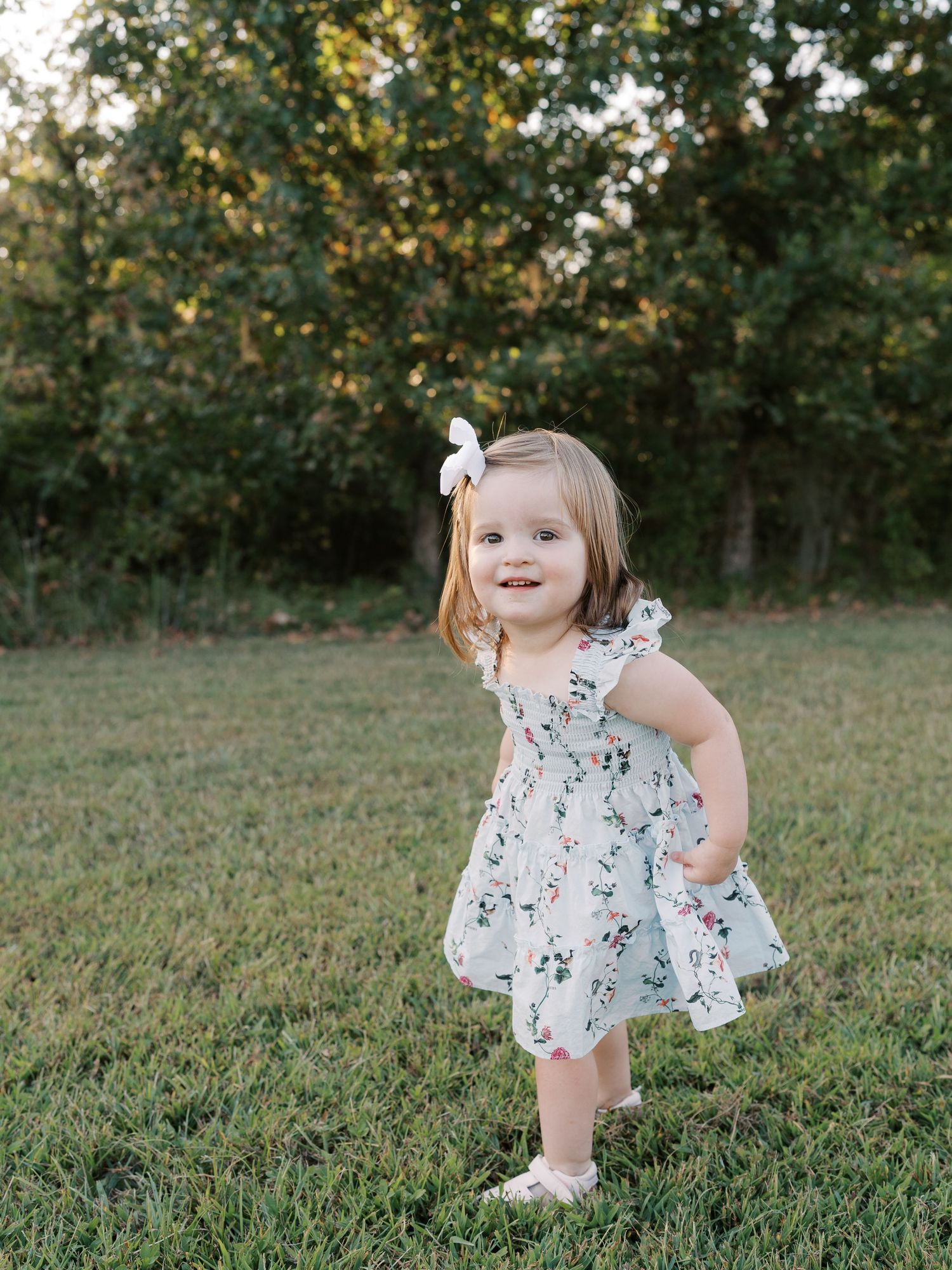 Toddler wearing white floral dress twirling and playing outdoors.