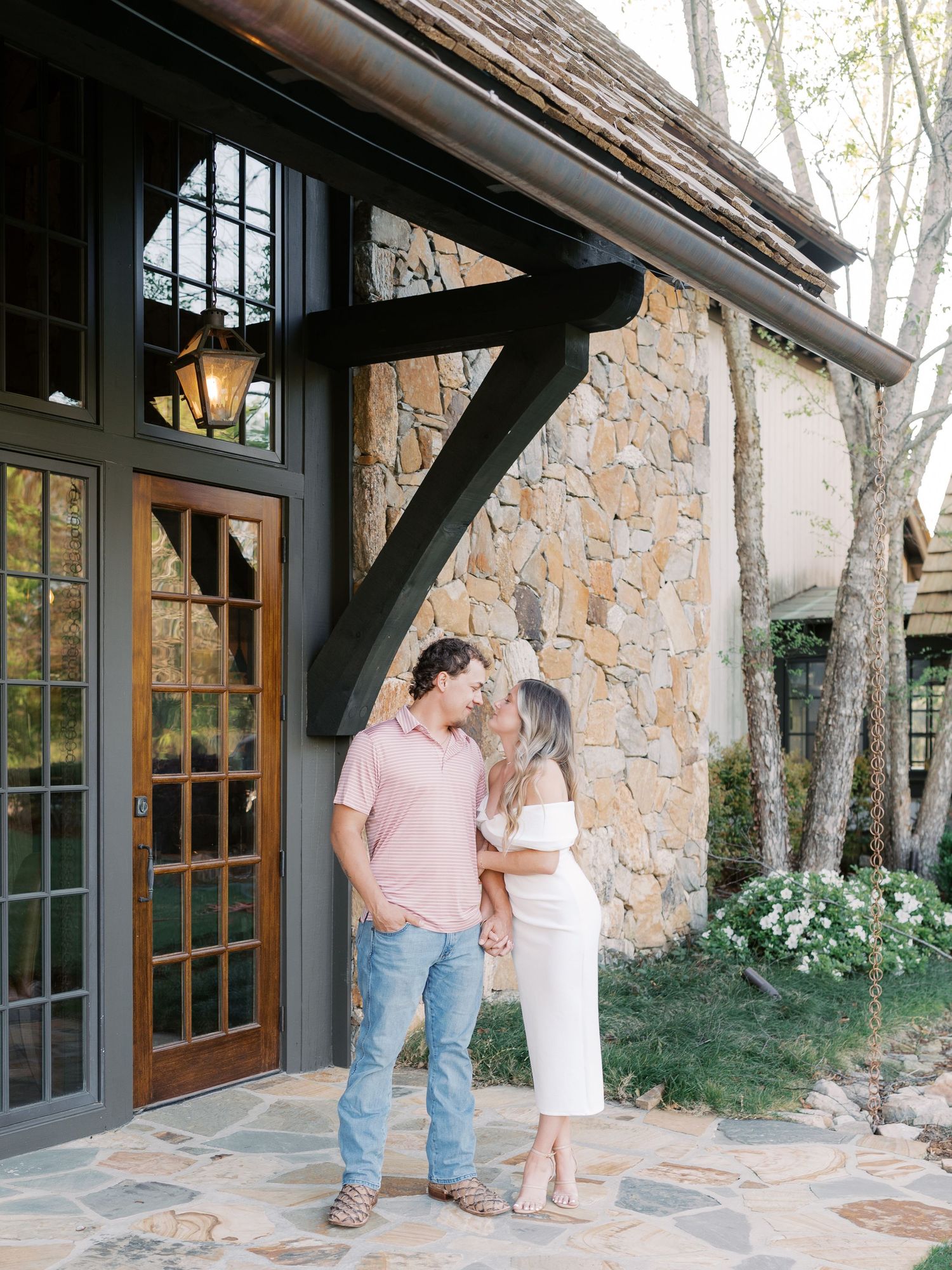 Couple stands together on stone patio outside rustic building with wooden doors.