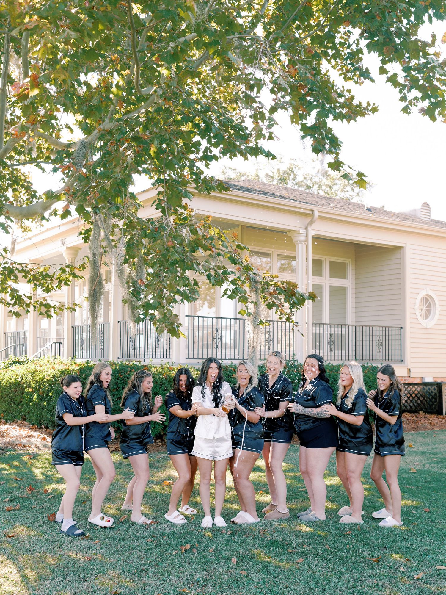 Bridal party in matching outfits posing outside white mansion under large tree.