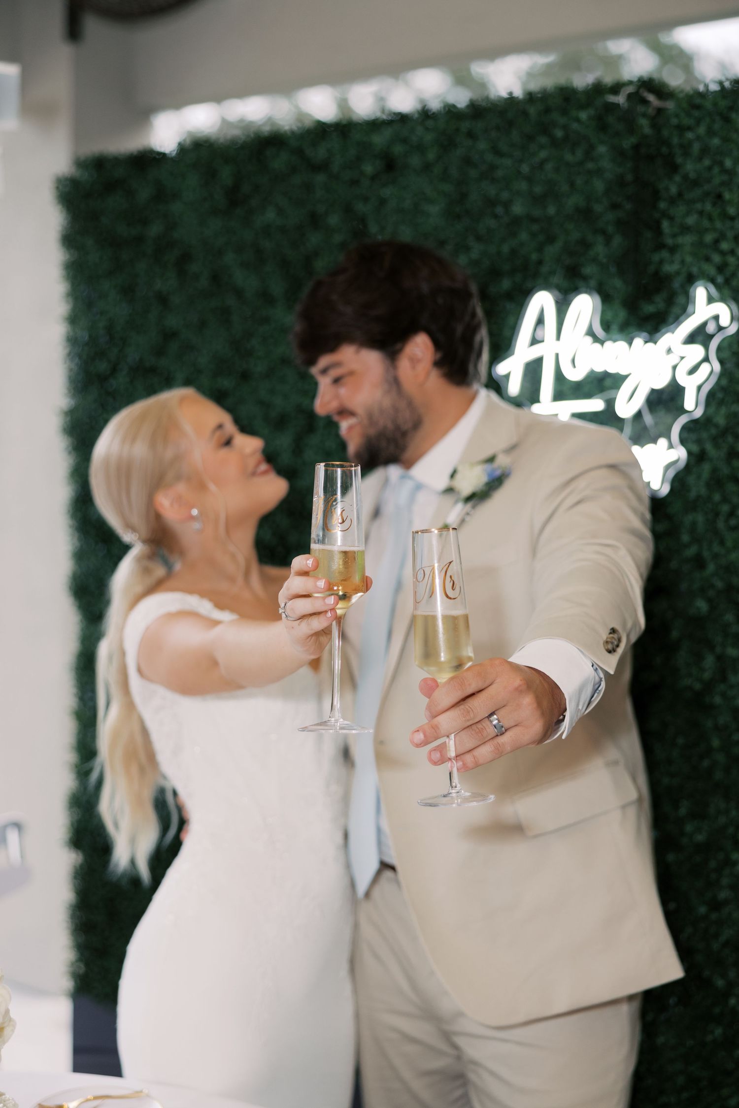 Couple sharing champagne toast in front of green hedge wall at wedding reception.