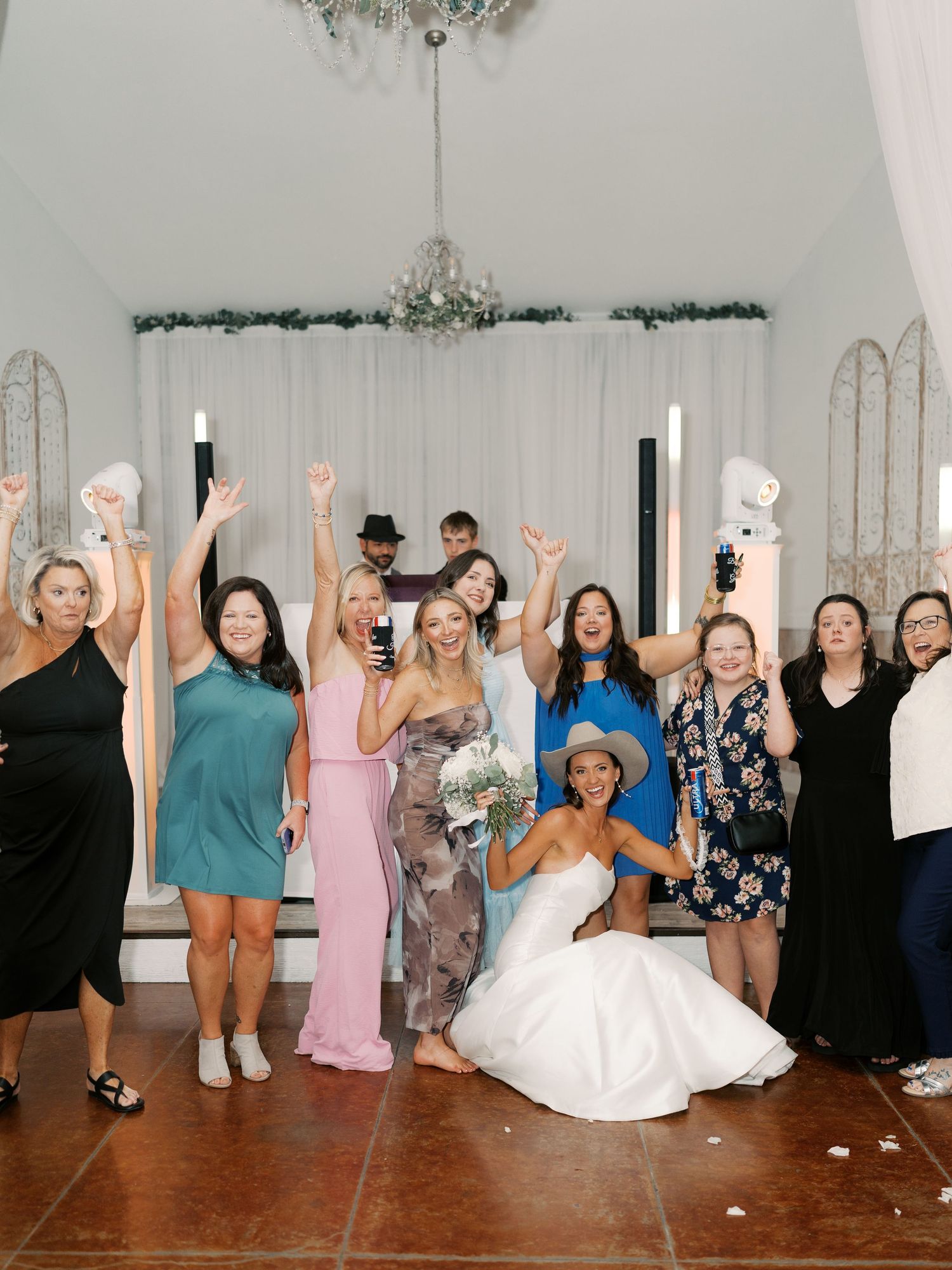 Wedding celebration group photo with guests raising drinks in a decorated white venue.