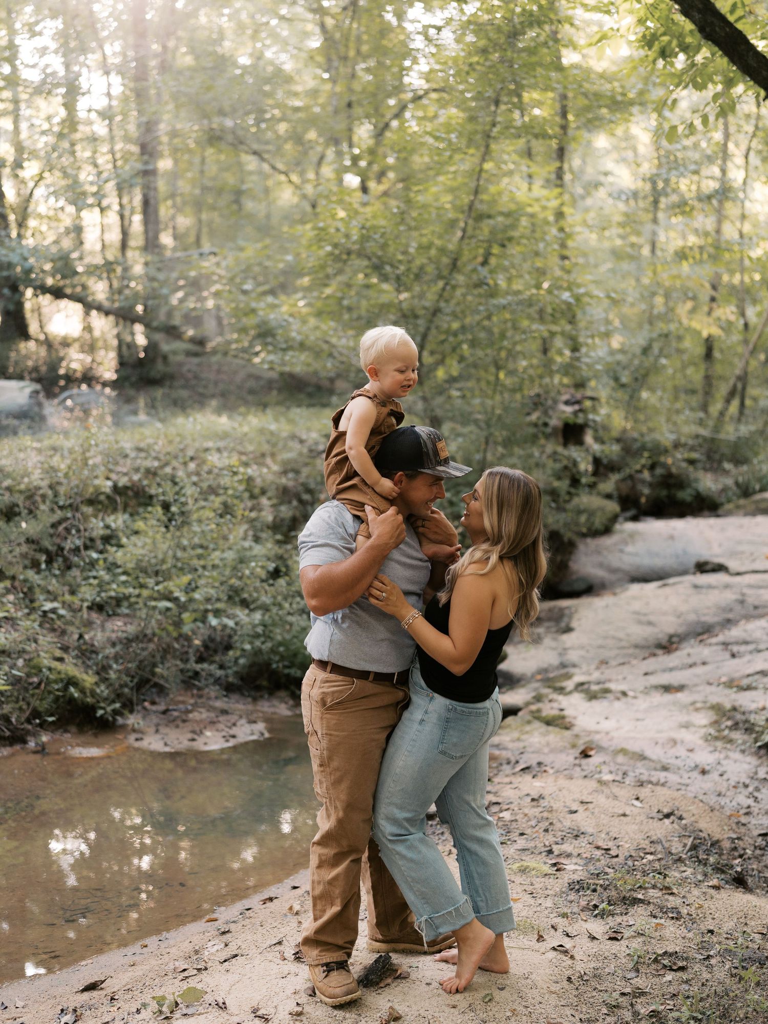 A family embraces while standing near a creek in a sunlit wooded area.