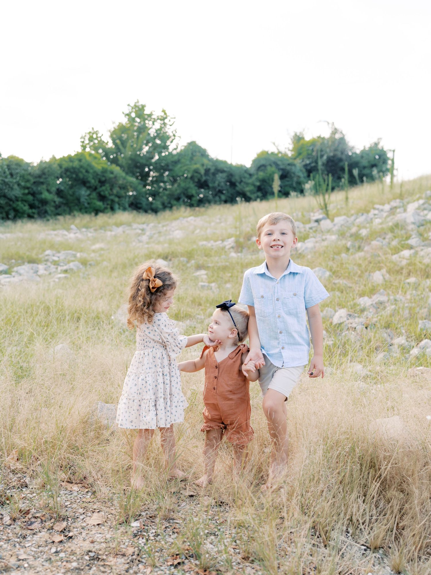 Three children playing together in a sunny field with rocks and tall grass.