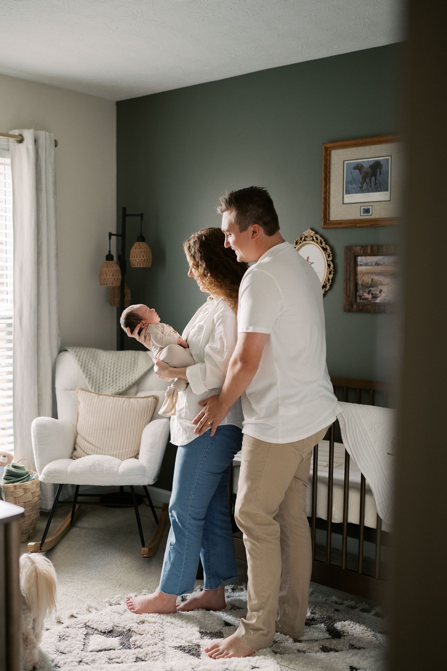 Family standing together in modern nursery with sage walls and white furniture.