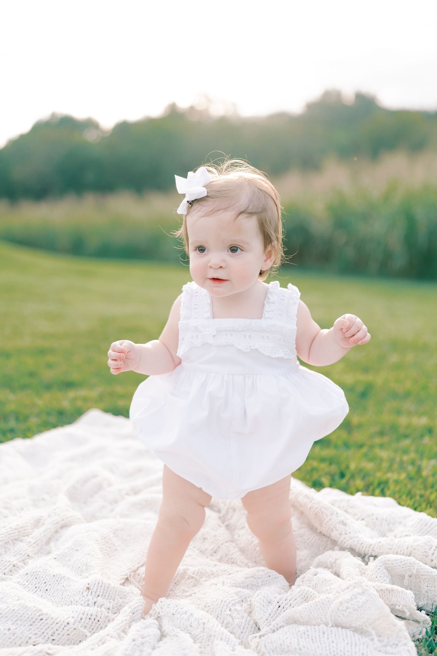 Baby wearing white dress sits on white blanket in sunny field.