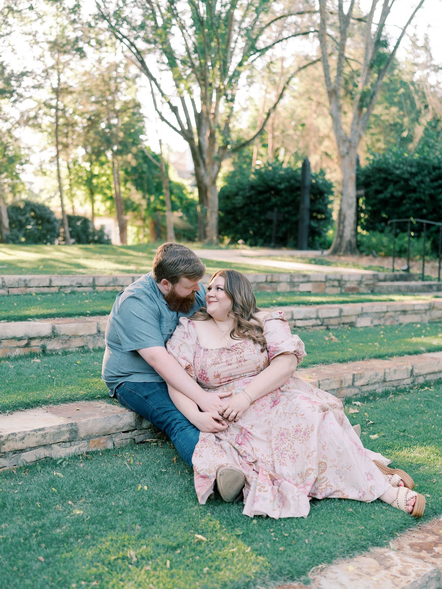 Romantic couple lounges on grass near stone wall in outdoor garden setting during sunset photoshoot.