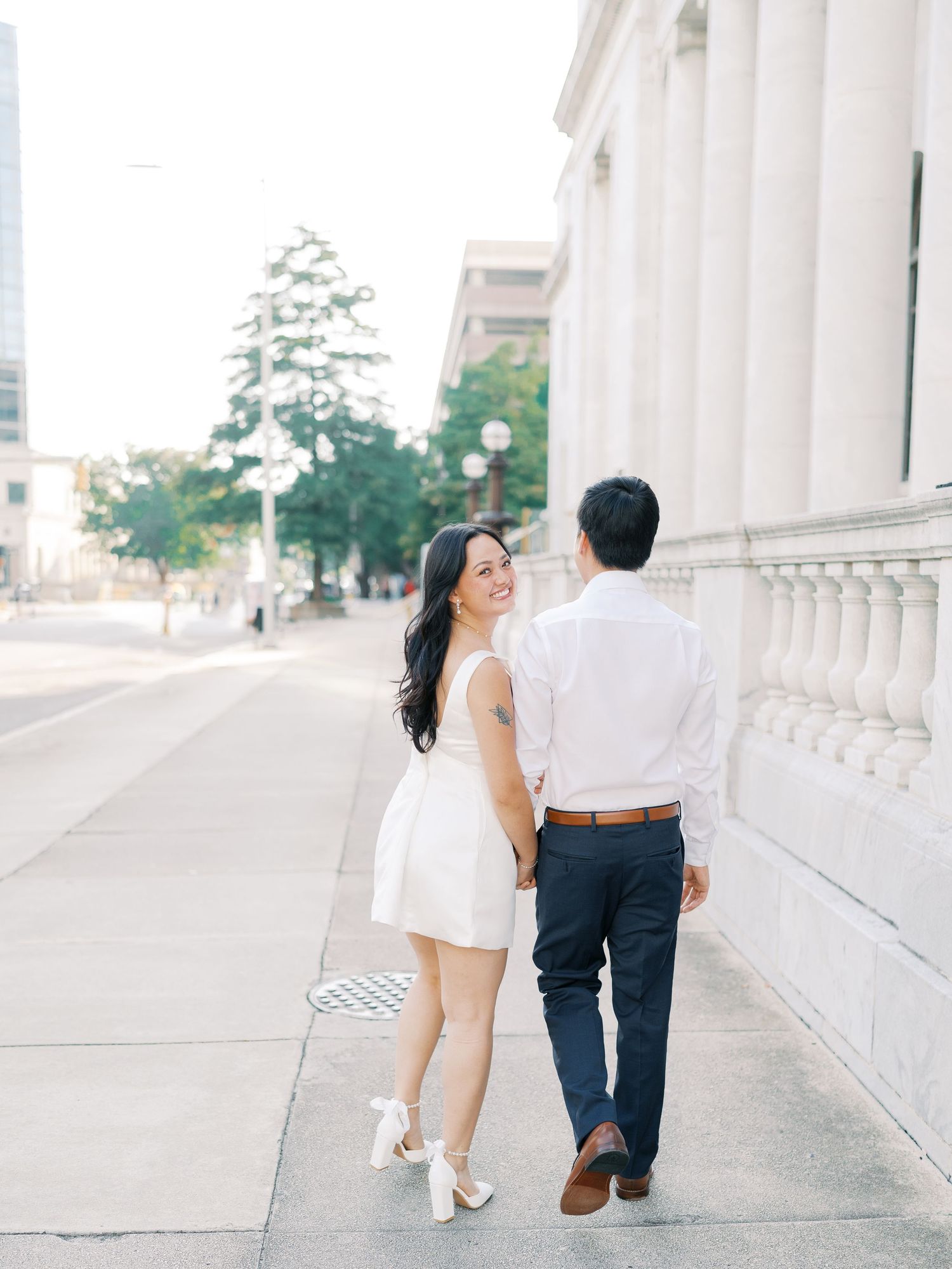 Romantic couple walking along elegant white columns in formal attire during sunset photo session.