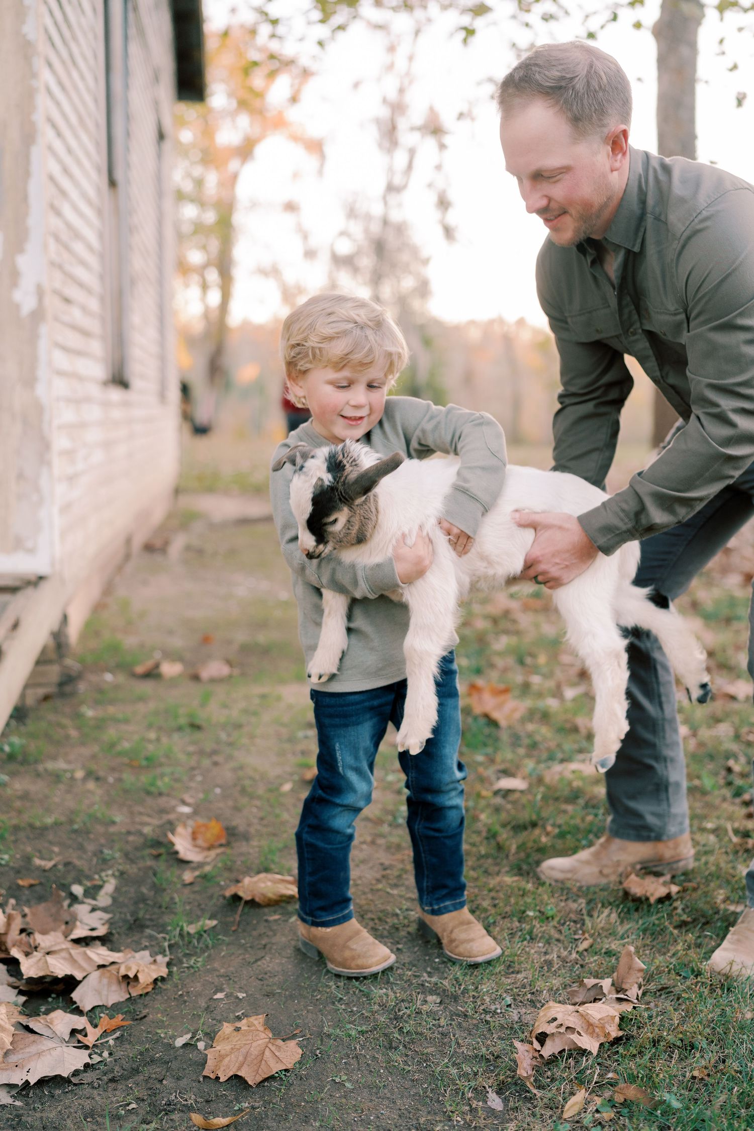 A child and adult play with a small white dog on fallen autumn leaves.