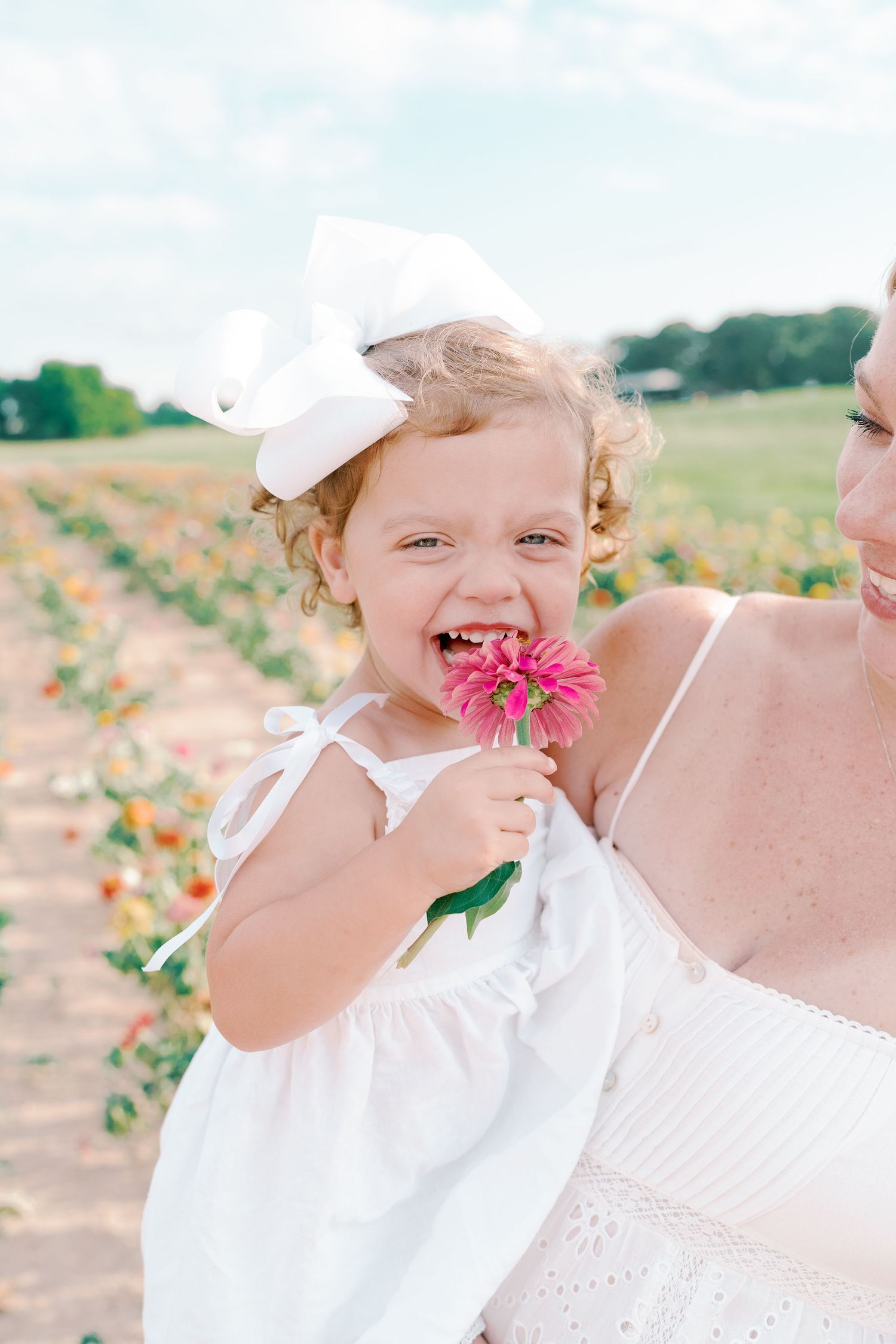Child in white dress smells pink flowers while playing in a summer flower field.