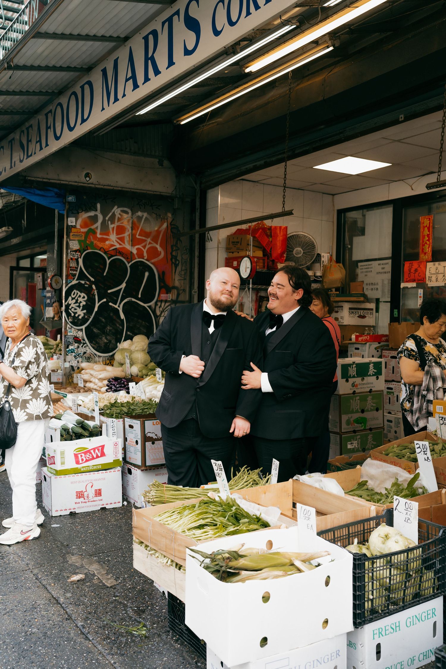 Customers browse fresh produce at an outdoor market stand with storefront signage.