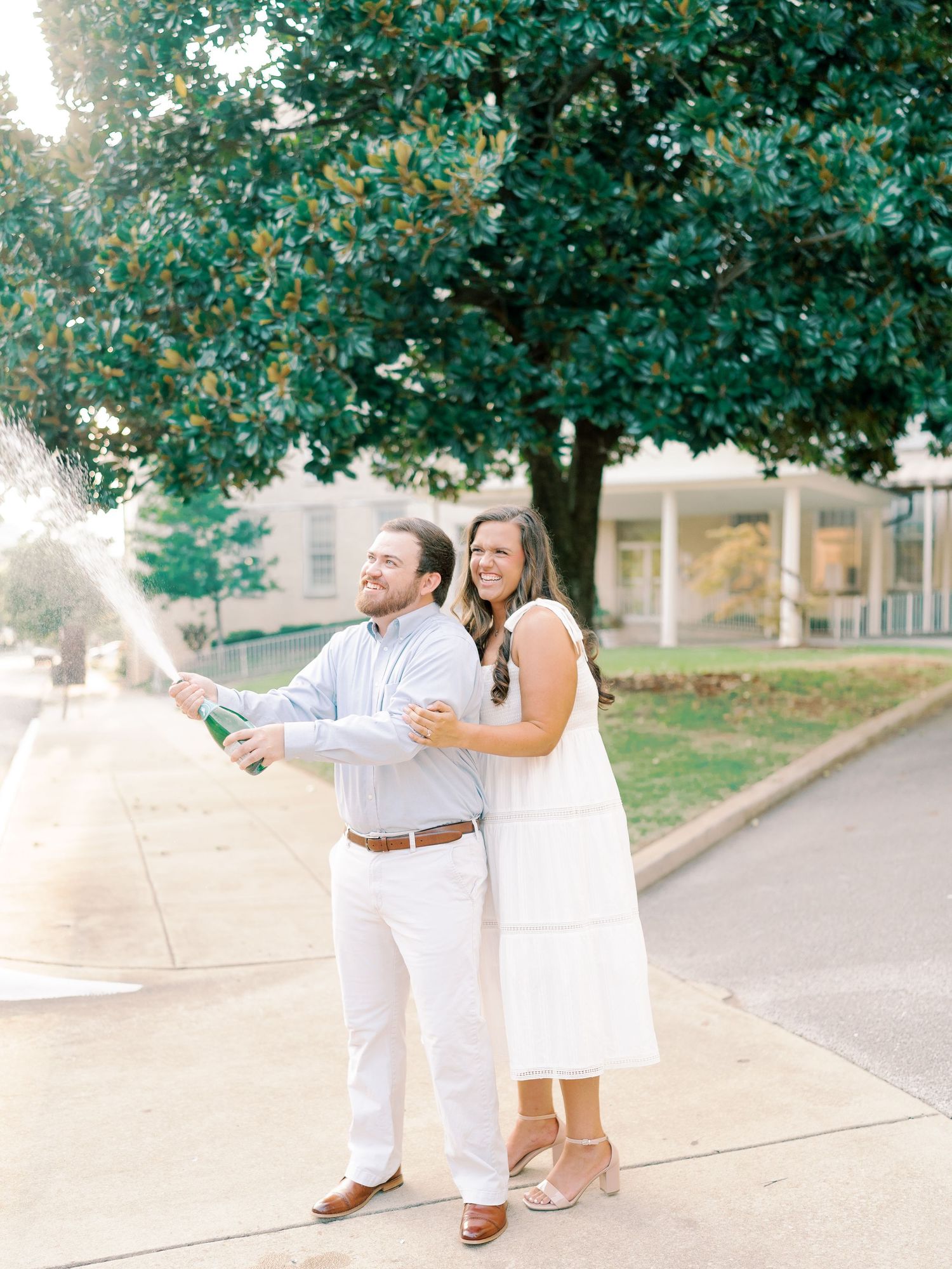 A couple in white outfits dancing playfully on a garden path surrounded by trees.