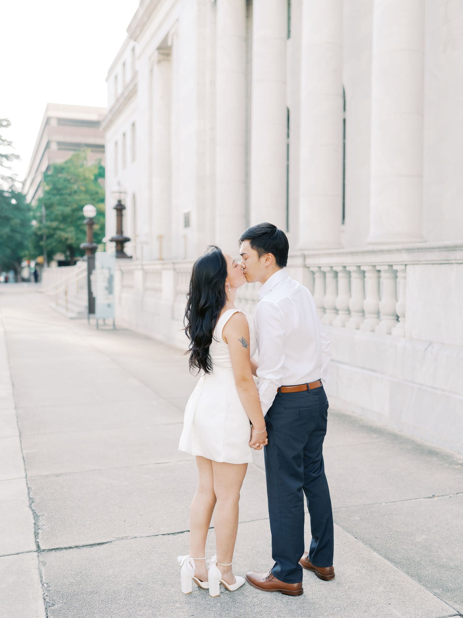 A couple in matching formal attire poses playfully by a white columned building exterior.