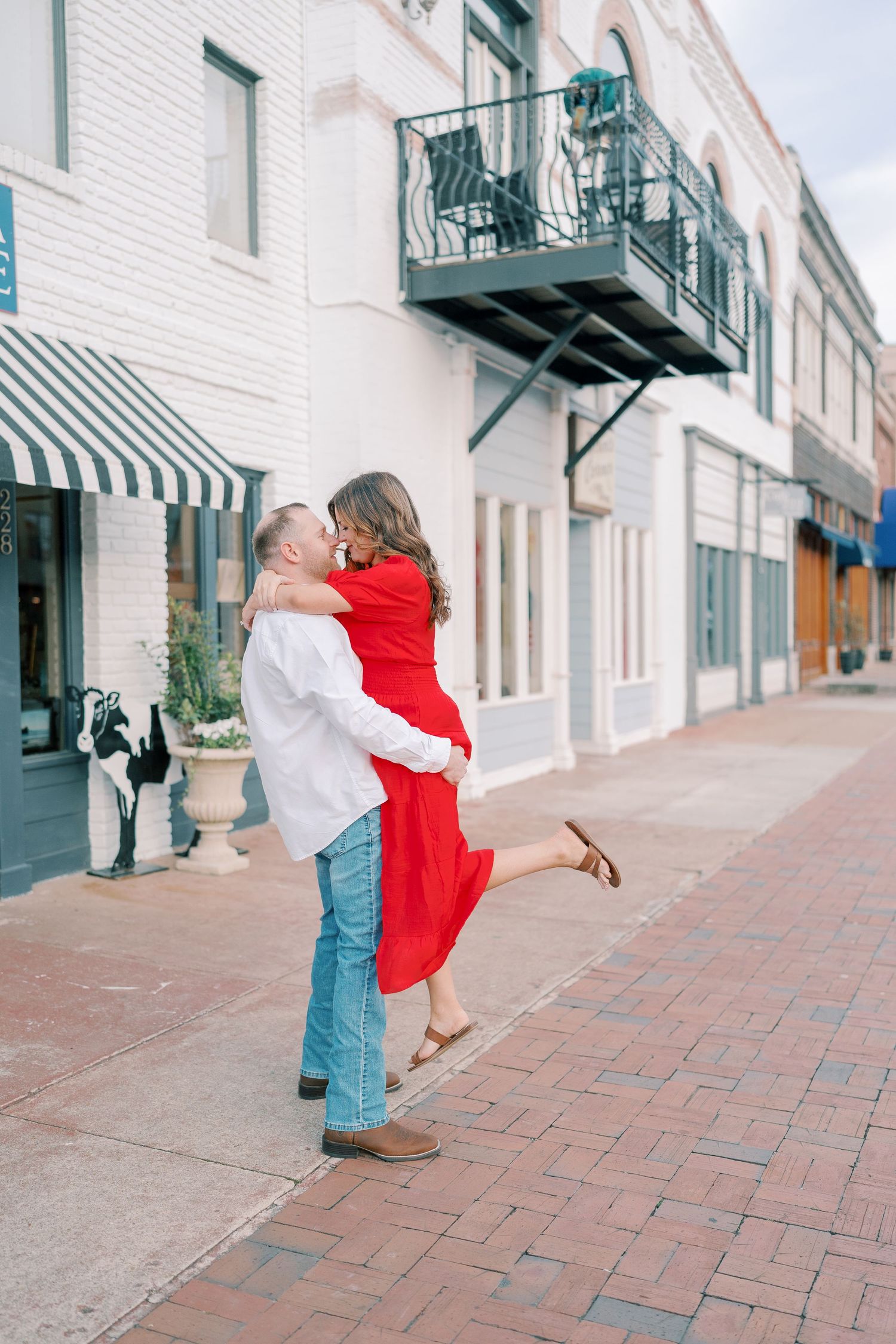 A couple in red dress and white shirt embrace romantically on a brick sidewalk outside historic buildings.