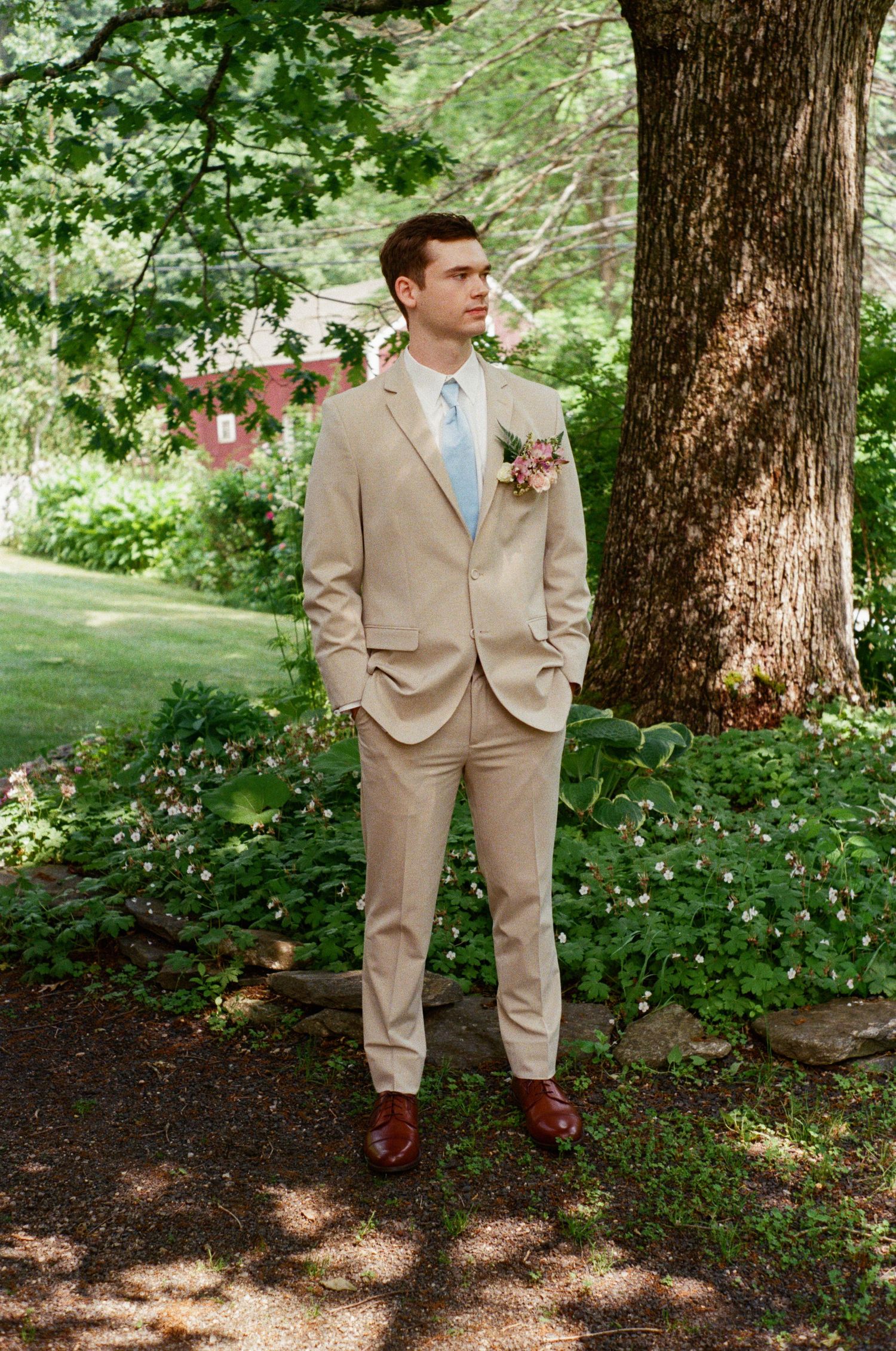 Groomsman in beige suit stands in dappled sunlight under large trees.