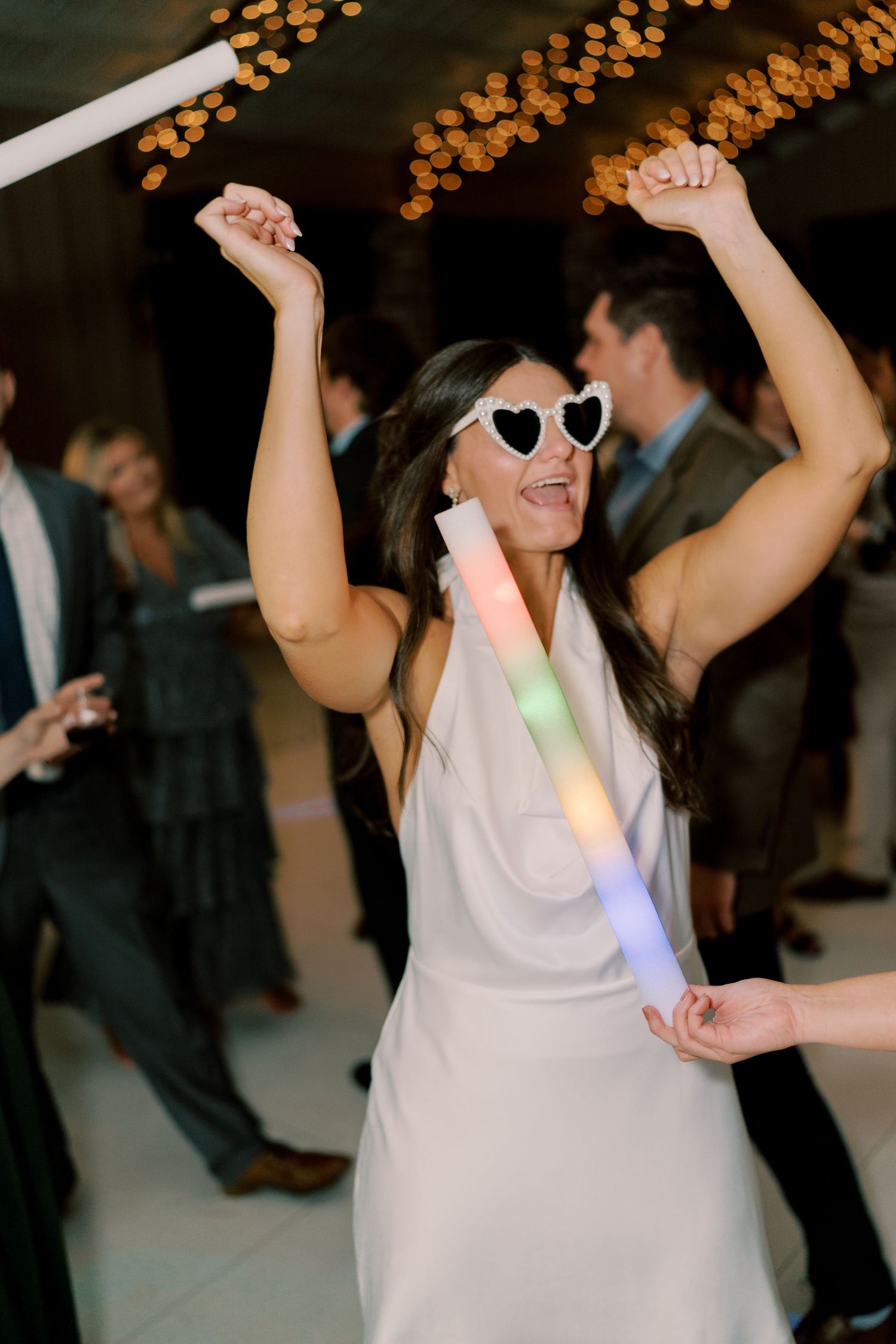 Party guest wearing heart-shaped sunglasses dances with arms raised under string lights.