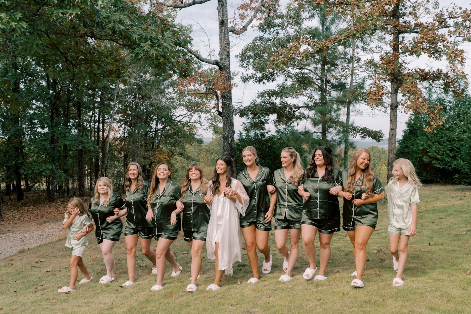 A playful outdoor wedding party poses together on a grassy hill with trees in the background.