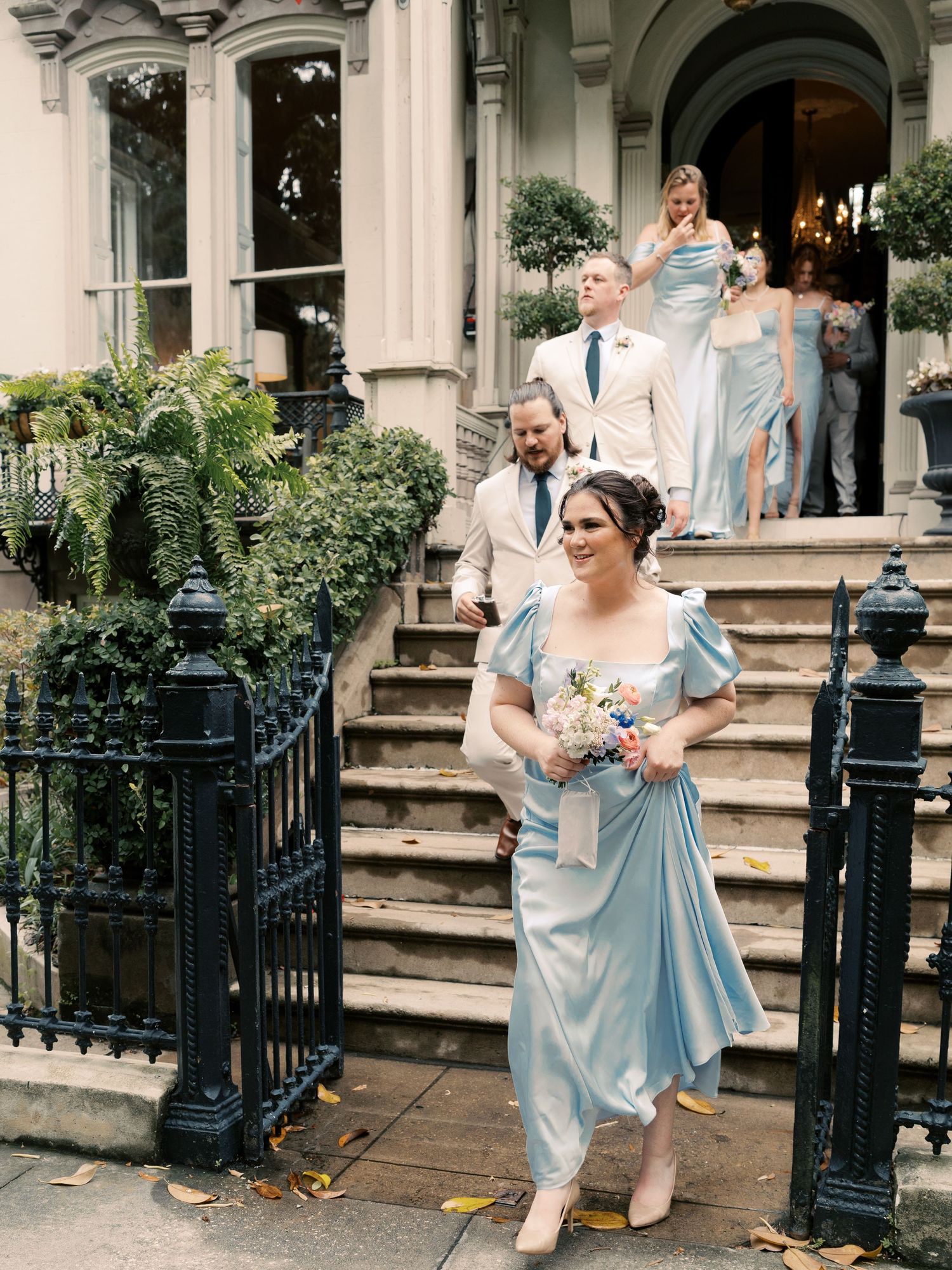 Wedding party in light blue dresses gathers on elegant stone steps of a historic building.