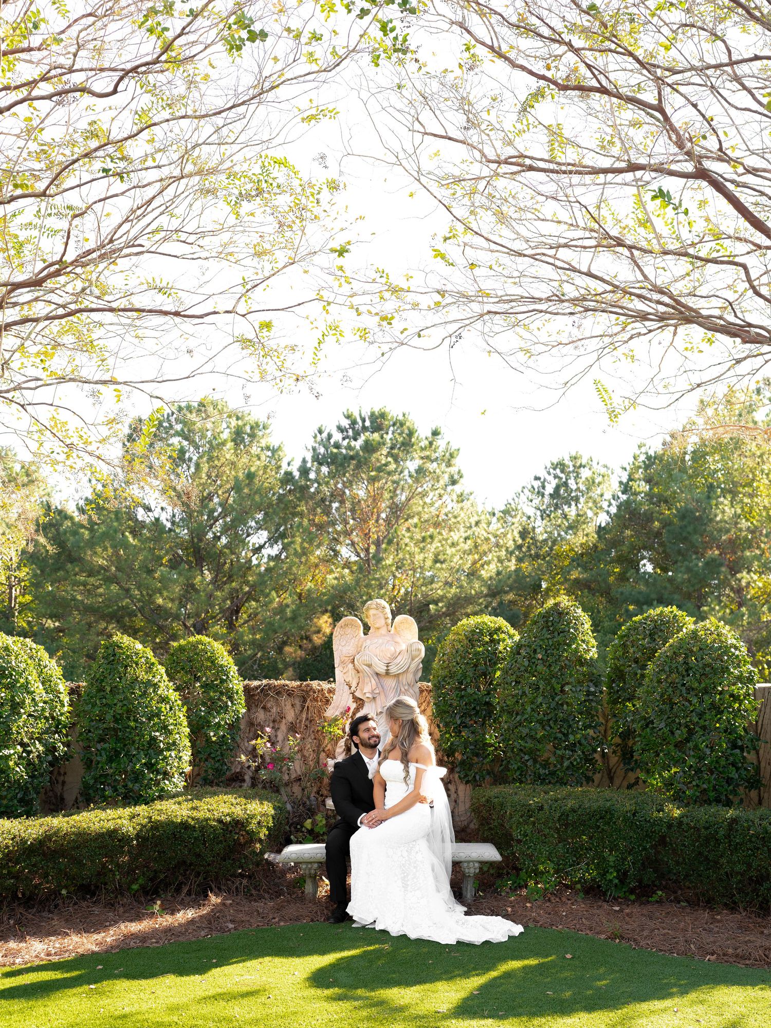 Newlyweds sit together on garden bench surrounded by manicured hedges and trees.